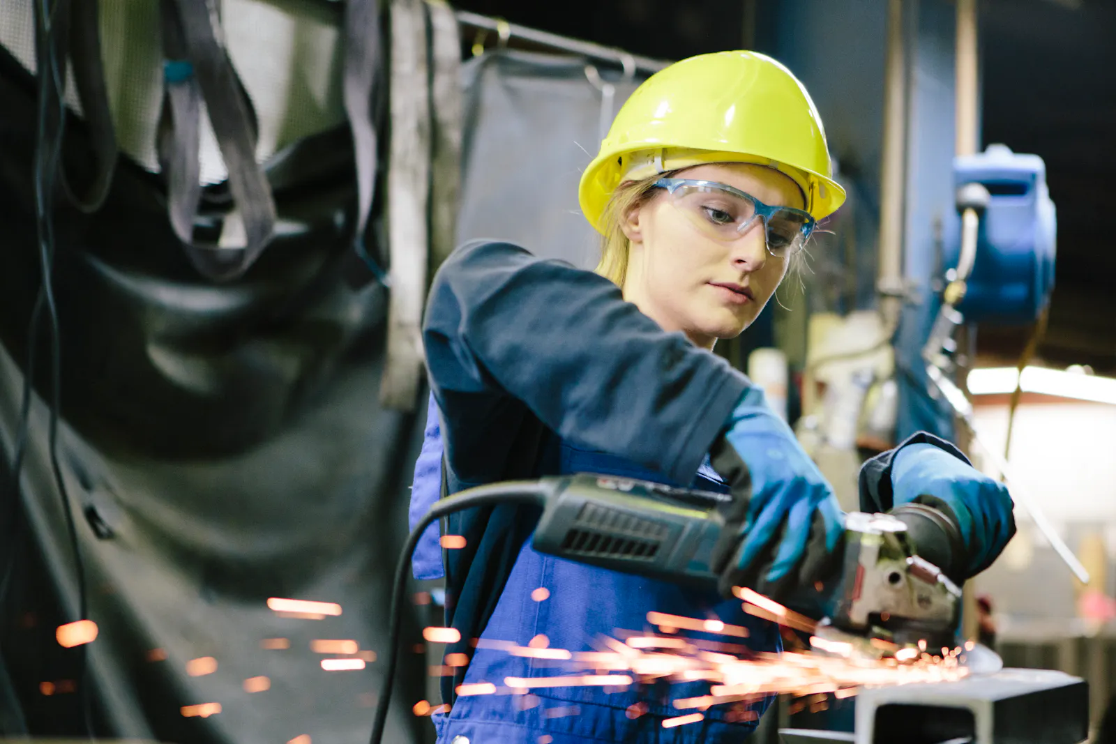 Das Bild zeigt eine junge Metallarbeiterin mit Schutzhelm und Schutzbrille. Sie schleift ein Metallteil mit dem Winkelschleifer; Funken sprühen in einer Werkhalle.