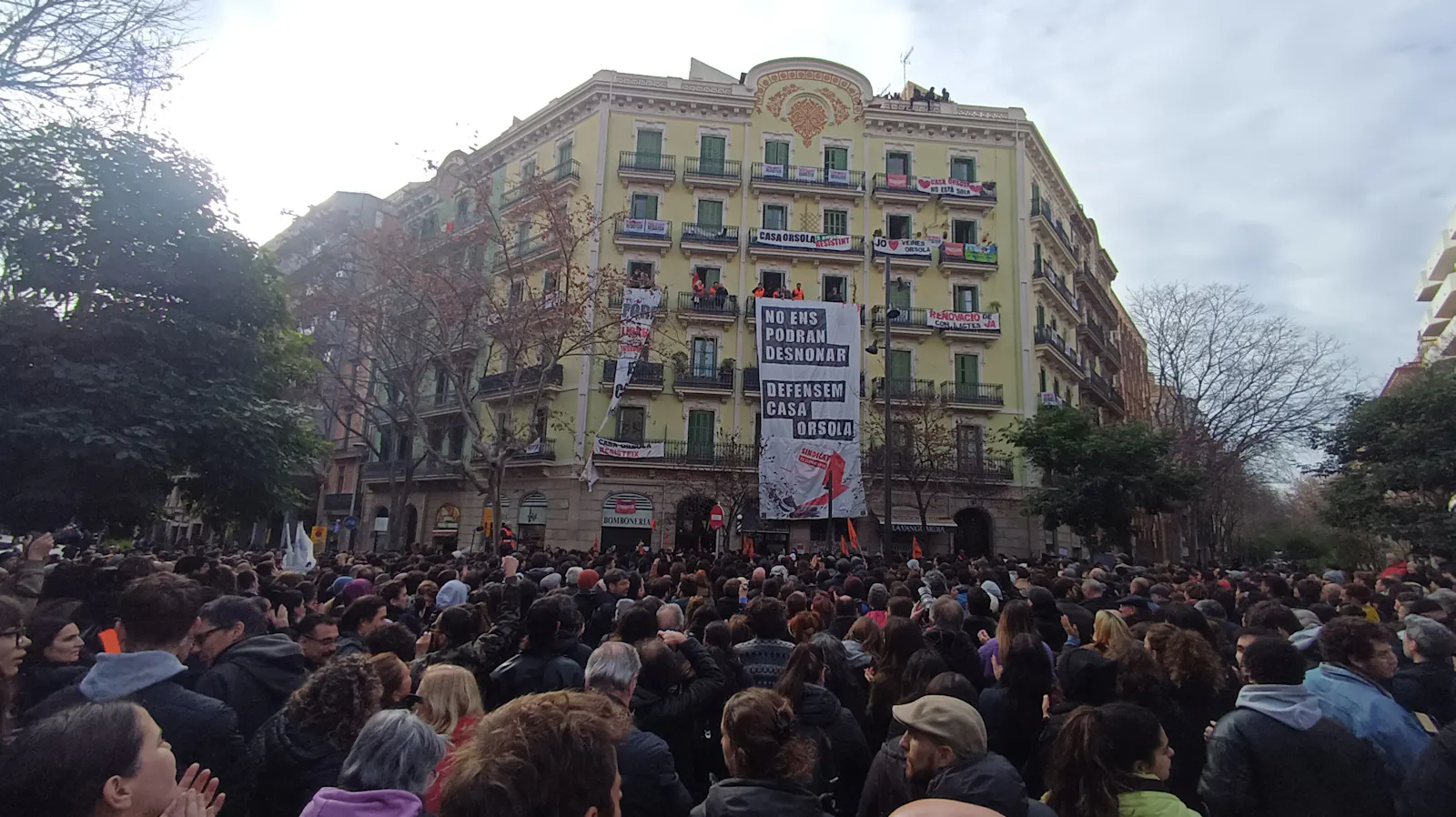 Ein hellgelb getünchter Jugendstilbau im großbürgerlichen Eixample in Barcelona. Vor Fassade hängt ein Protestplakat. Vor dem Gebäude stehen Tausende Demonstranten.