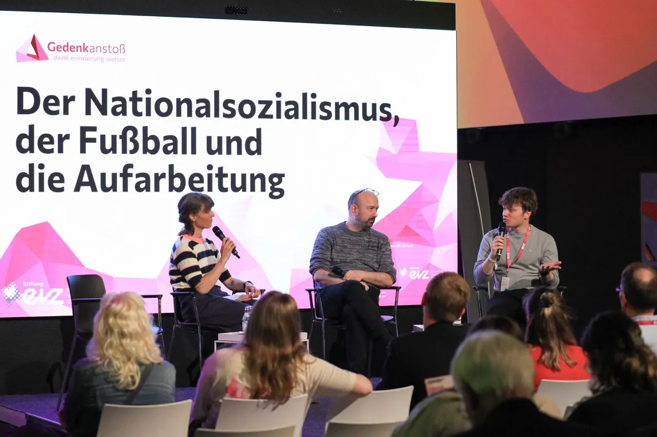 Auf einer Bühne sitzen drei Menschen: Die Journalistin Nora Hespers, die ein Mikrofon vor ihren Mund hält, EVZ-Projektleiter Jens Schley und Leon Walter, der an der MEMO-Studie als Wissenschaftlter mitgearbeitet hat. Im Hintergrund ist auf einer LED-Leinwand das Logo Gedenkanstoß zu sehen. Darunter in großen schwarzen Buchstaben: Der Nationalsozialismus, der Fußball und die Aufarbeitung.