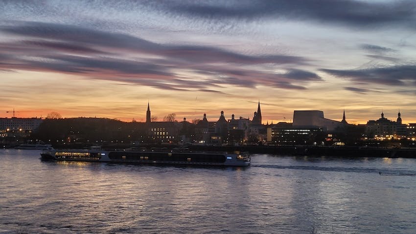 Blick auf das Stadtpanorama von Bonn von der Beueler Uferseite aus, aufgenommen bei Sonnenuntergang, im Vordergrund der Rhein mit einem Flusskreuzfahrtschiff. Foto: Christine Piontek