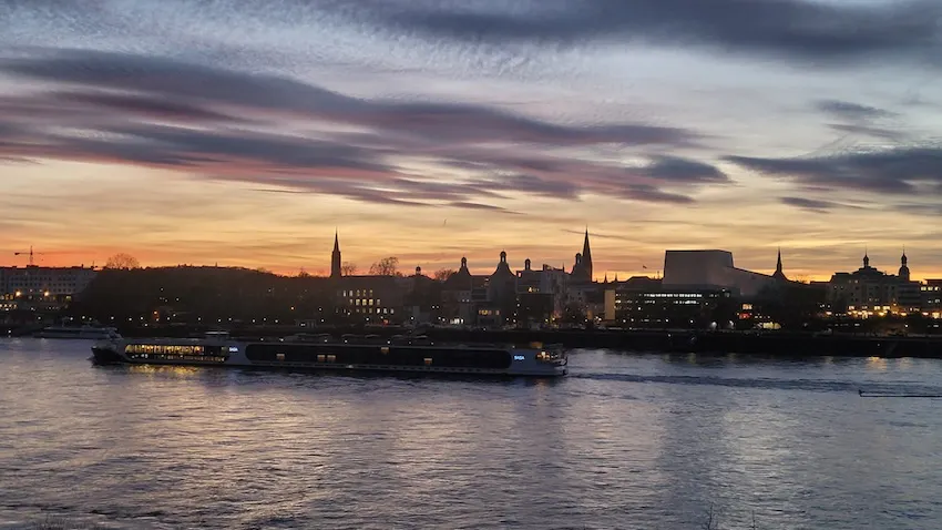 Blick auf das Stadtpanorama von Bonn von der Beueler Uferseite aus, aufgenommen bei Sonnenuntergang, im Vordergrund der Rhein mit einem Flusskreuzfahrtschiff. Foto: Christine Piontek