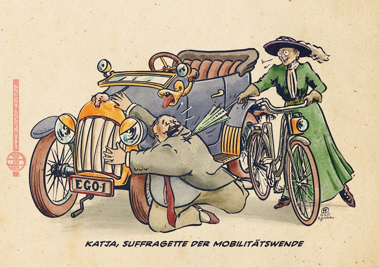 Karikatur von Guido Kühn (guidos-welt.de): Eine Frau im edwardianischen Kostüm – grünes Kleid, großer Hut, Brille – schiebt lächelnd ihr Fahrrad an einem wild um sich schlagenden Mann an einem Oldtimer vorbei. Das Auto mit dem Kennzeichen „EGO-1" hat ein aufgerissenes Gesicht und streckt die Zunge heraus. Unterschrift: „Katja, Suffragette der Mobilitätswende."