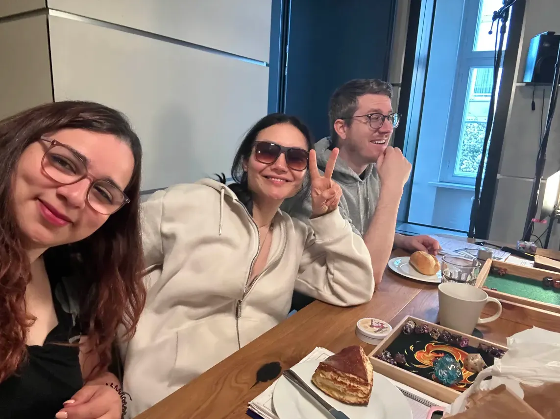 Aylin, Rubi und Tim sitzen nebeneinander und lächeln. Vor ihnen stehen Tassen und Teller mit Kaffee und Brötchen.
