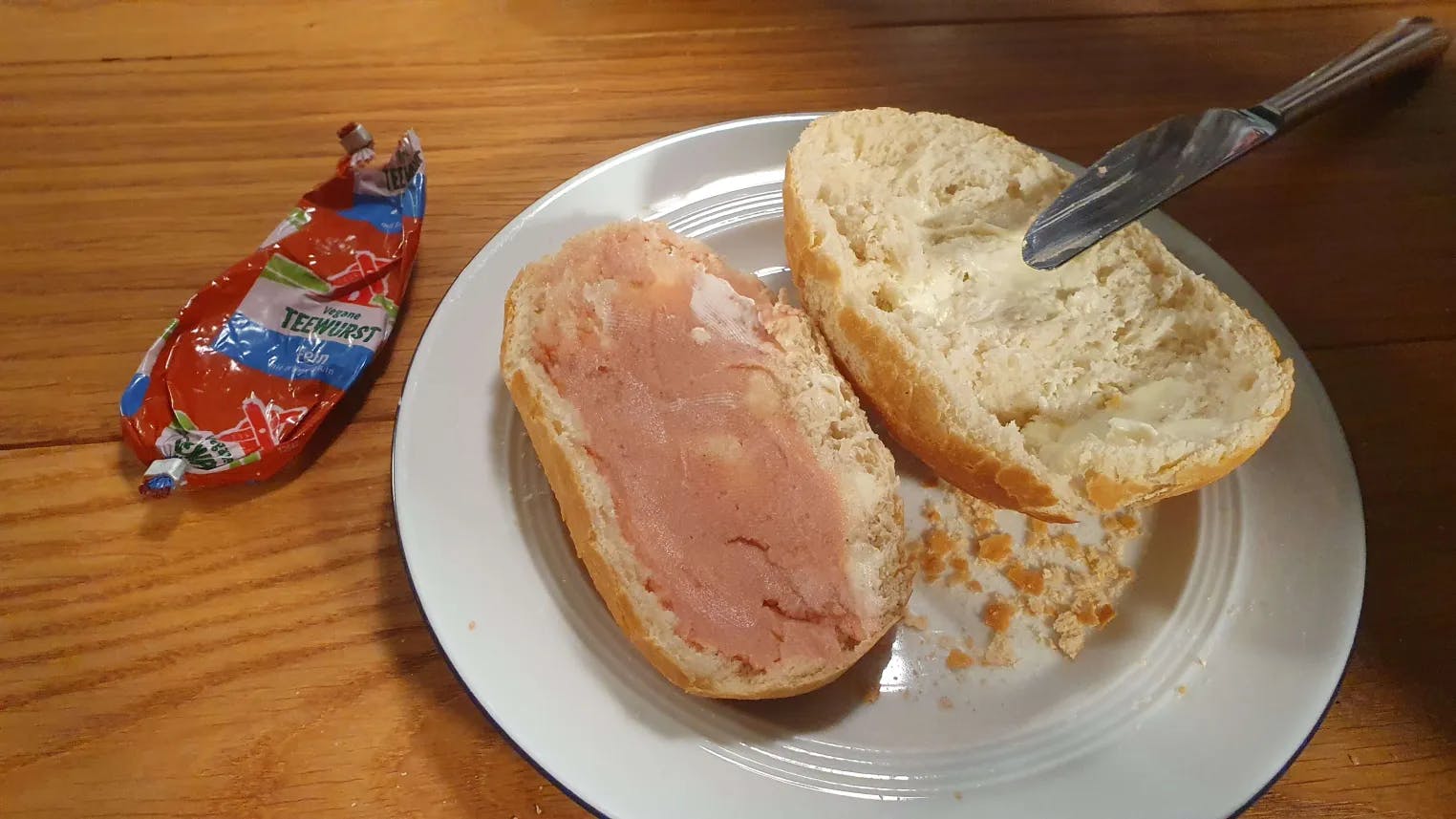 Ein Bild von einem aufgeschnittenen Brötchen, die eine Hälfte ist beschmiert mit Teewurst -  daneben liegt die vegane Teewurstpackung.