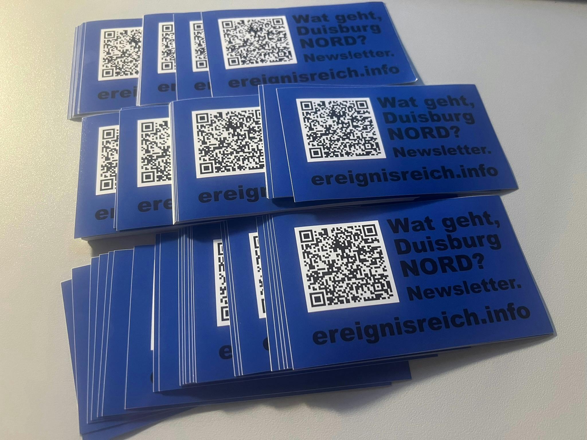 Mehrere Stapel rechteckiger blauer Aufkleber mit einem QR-Quode und der Aufschrift: Wat geht Duisburg Nord? Newsletter. Ereingnisreich.info