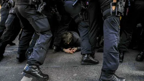 Demonstrant am Boden, überall Polizist:innen, von denen nur Beine, Stiefel und die Gürtel mit Waffen etc. zu sehen sind