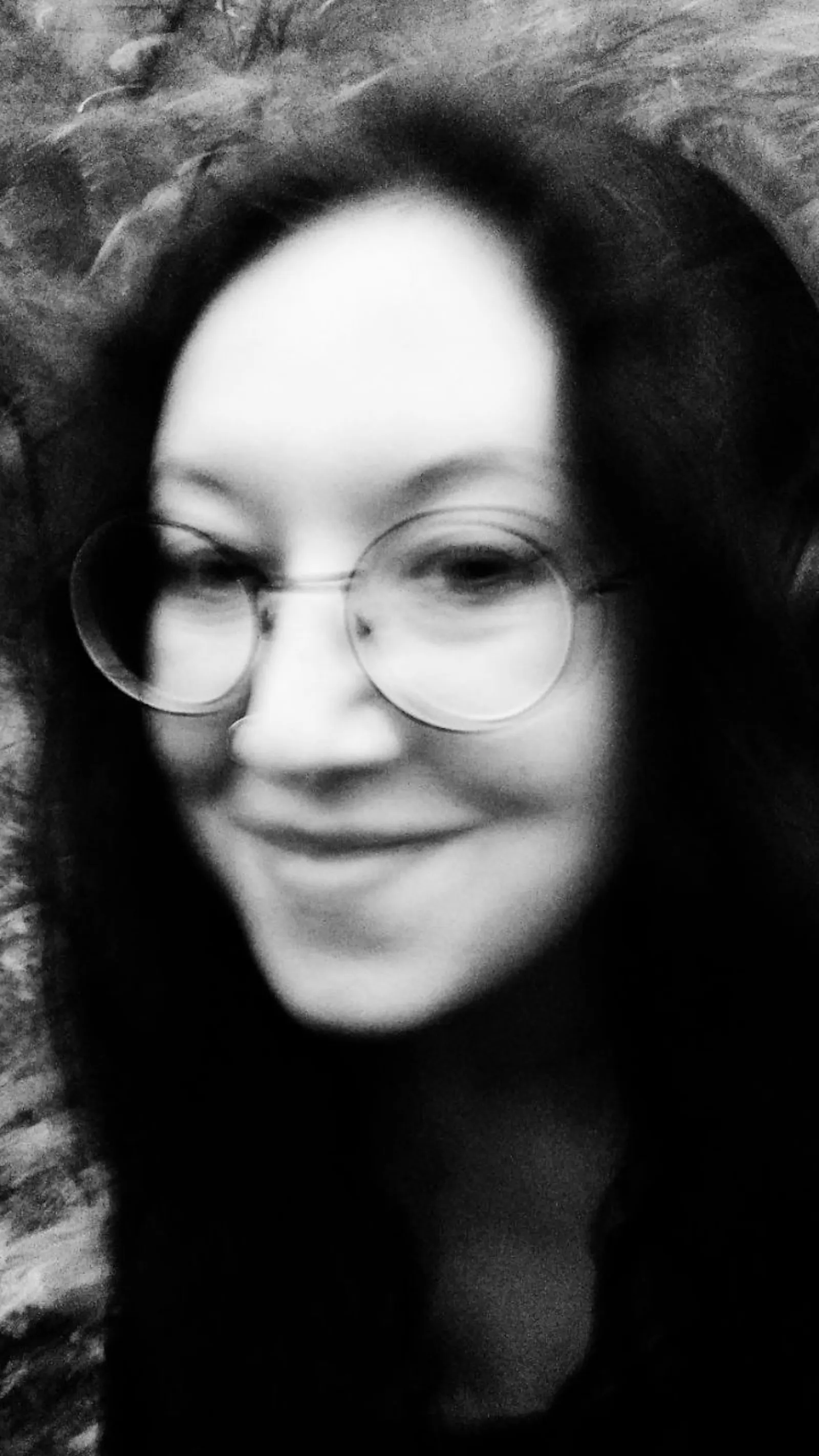 Schwarz-weiß Fotografie, verwackelt. Eine Frau lächelt in die Kamera, sie trägt eine Brille und die dunklen Haare offen. Im Hintergrund sind Bäume und Sträucher.