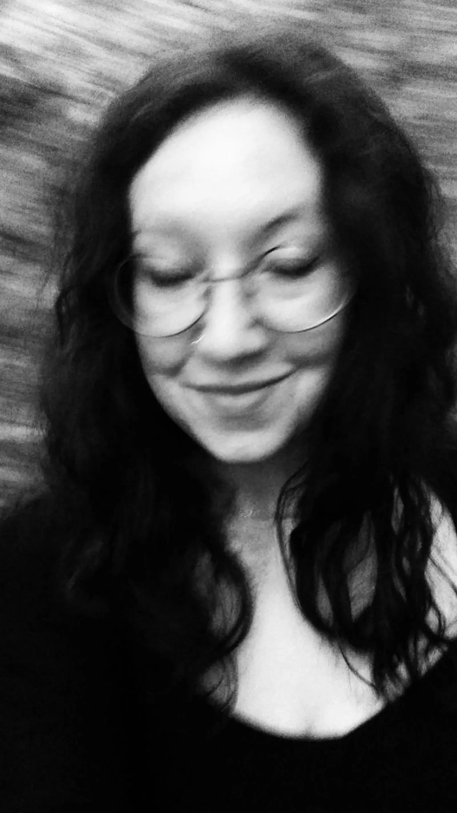 Schwarz-weiß Fotografie, verwackelt. Eine Frau senkt den Blick, sie trägt eine Brille und die dunklen Haare offen. Im Hintergrund sind Bäume und Sträucher.