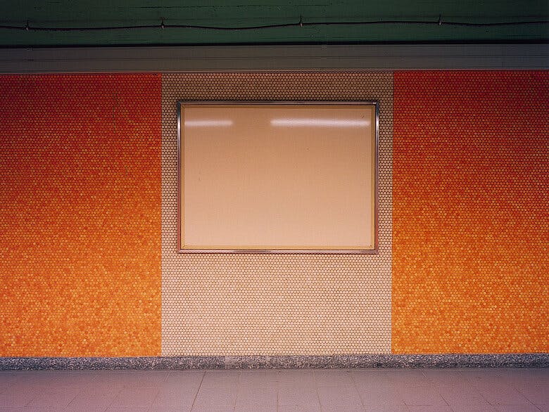 Foto von Götz Diergarten. Zeigt eine orangene Wand mit weißer Plakatfläche in einer Brüsseler Ubahn Station.