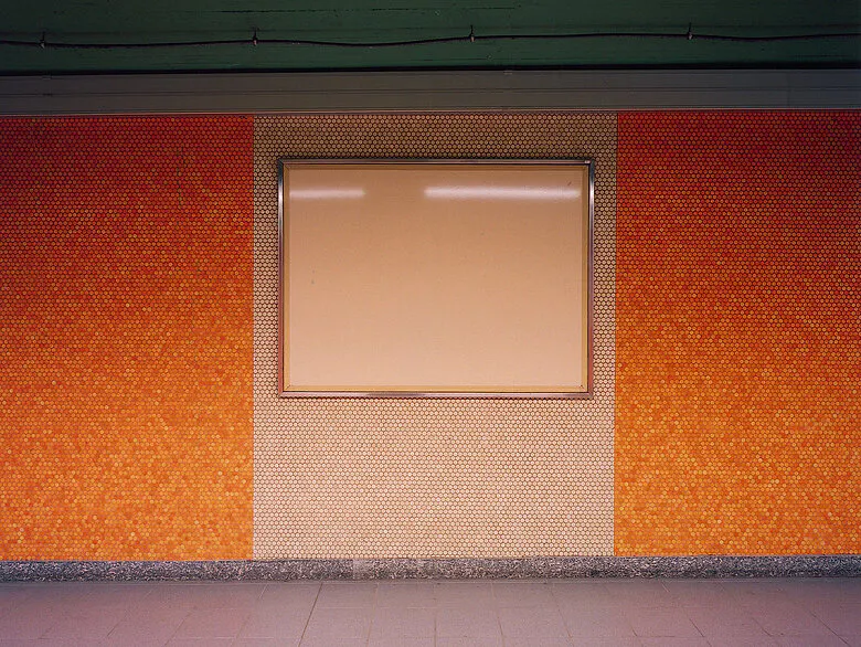 Foto von Götz Diergarten. Zeigt eine orangene Wand mit weißer Plakatfläche in einer Brüsseler Ubahn Station.
