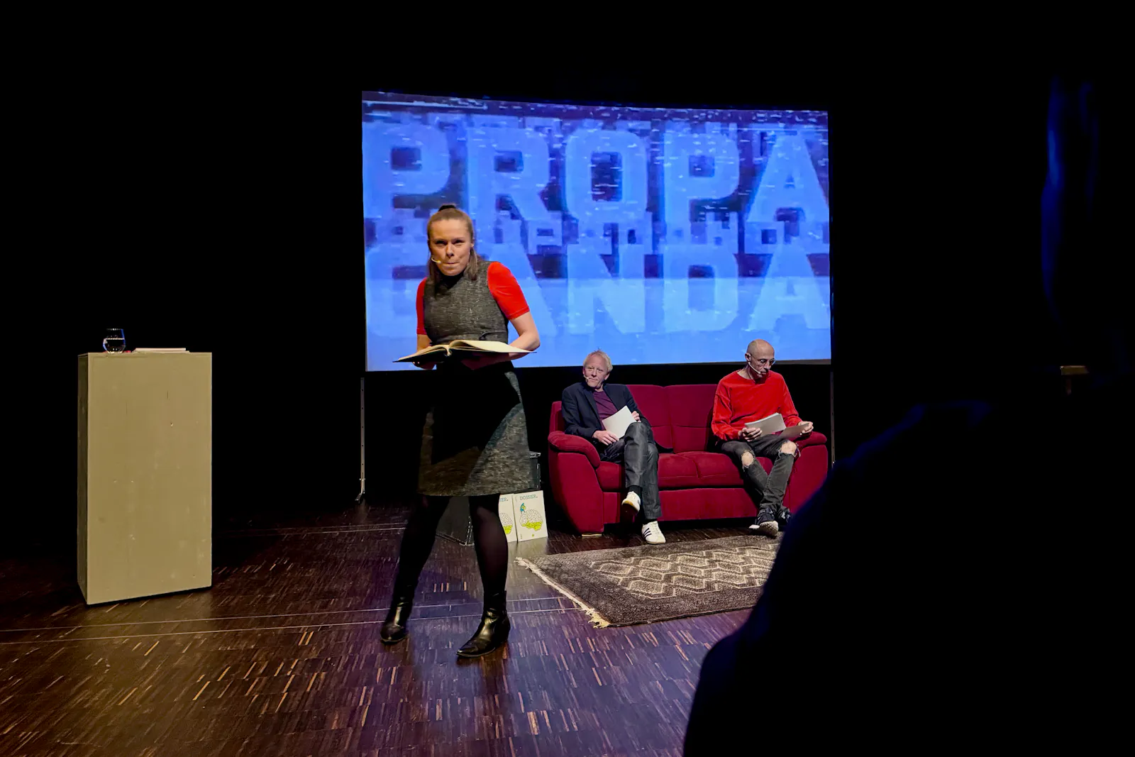 Schauspielerin vor Bildschirm mit Propanda-Schriftzug und Sofa mit zwei Männern