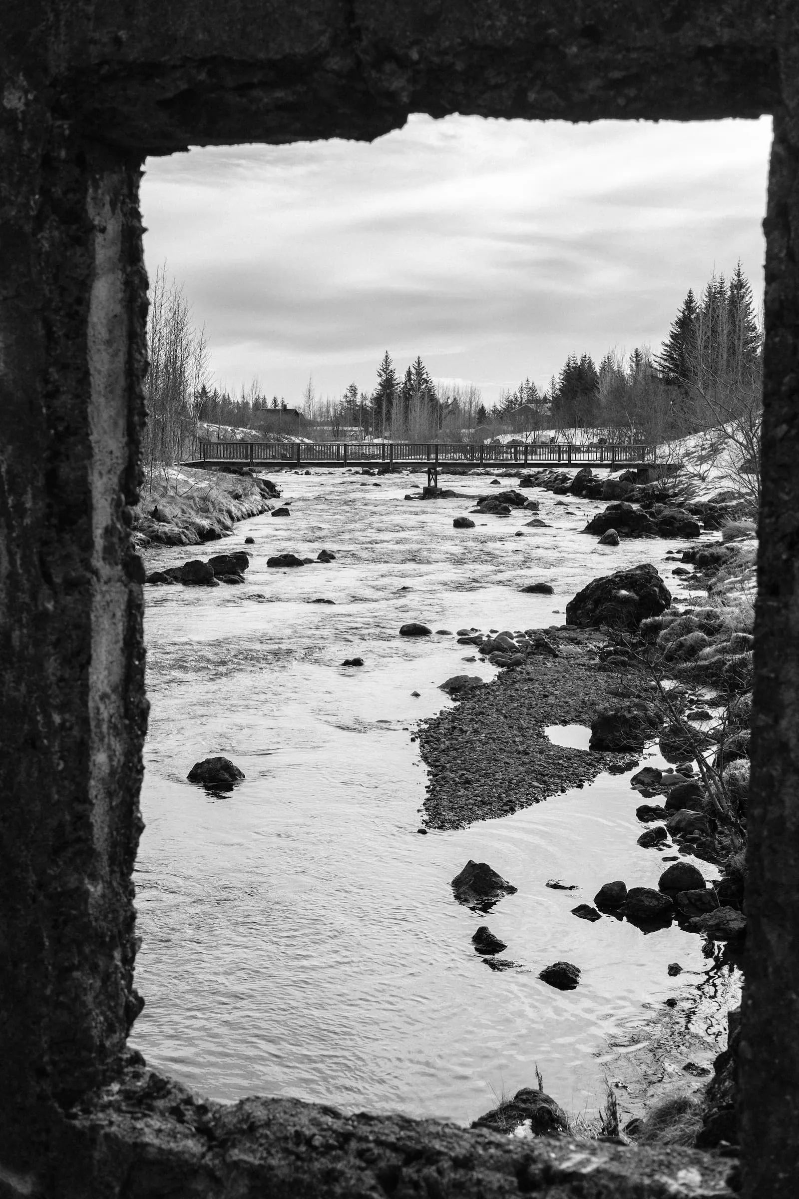 Through a ruined wall, we see a pedestrian bridge over a river