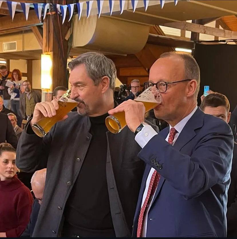 Markus Söder und Friedrich Merz trinken Hefeweizen in einem Festsaal