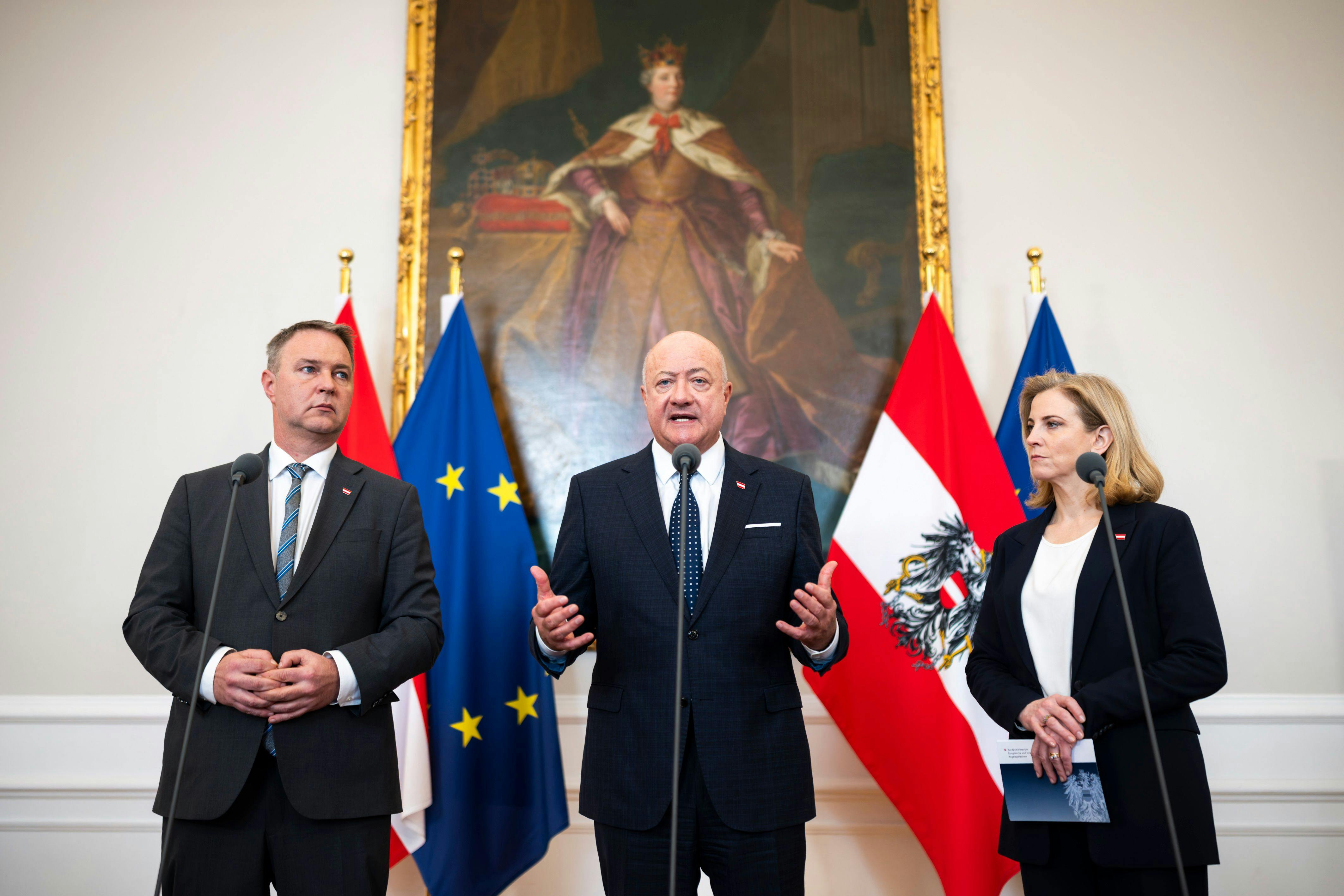 Das Foto zeigt Bundeskanzler Christian Stocker, Vizekanzler Andreas Babler und Außenministerin Beate Meinl-Reisinger. Sie stehen nebeneinander vor Mikrofonen und österreichischen sowie EU-Fahnen in einem repräsentativen Saal des Bundeskanzleramts. Im Hintergrund hängt ein großes Gemälde.