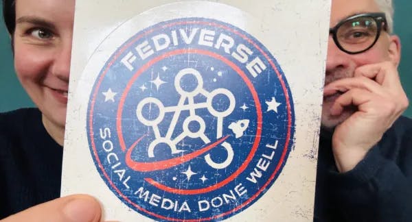 [Ilona und Marek halten einen Aufkleber hoch. Auf dem steht "FEDIVERSE - SOCIAL MEDIA DONE WELL"]