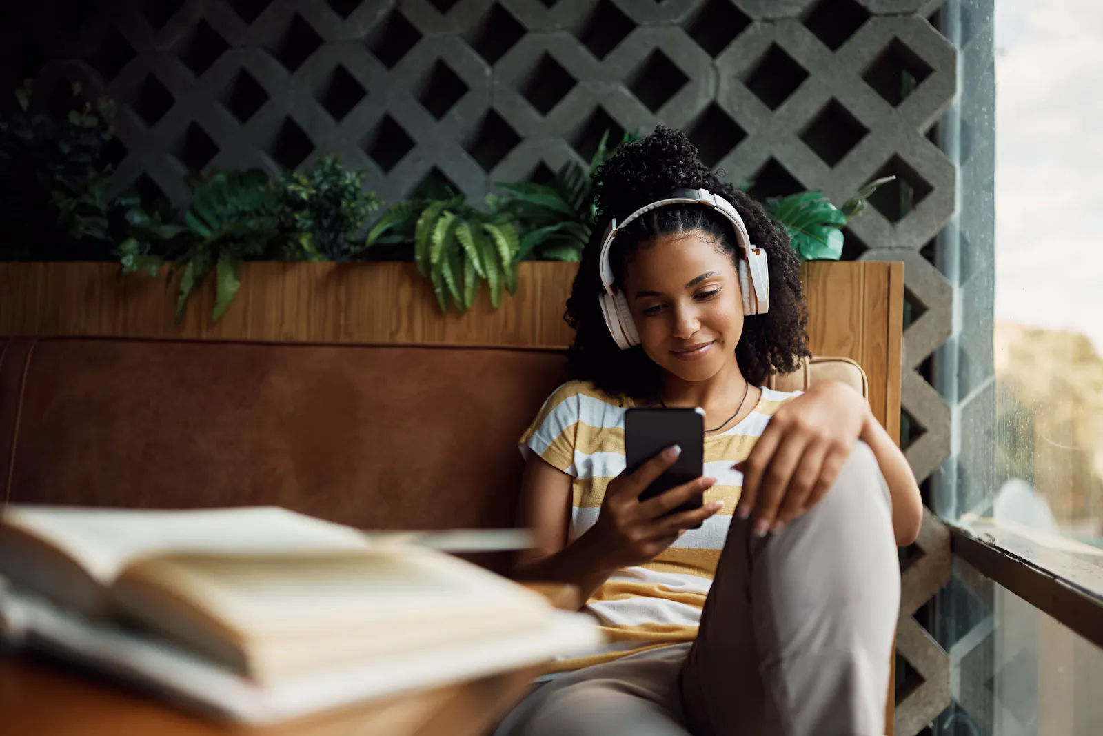 A beautiful black woman wearing white headphones and looking at her phone in a brightly lit room with some plants behind her.