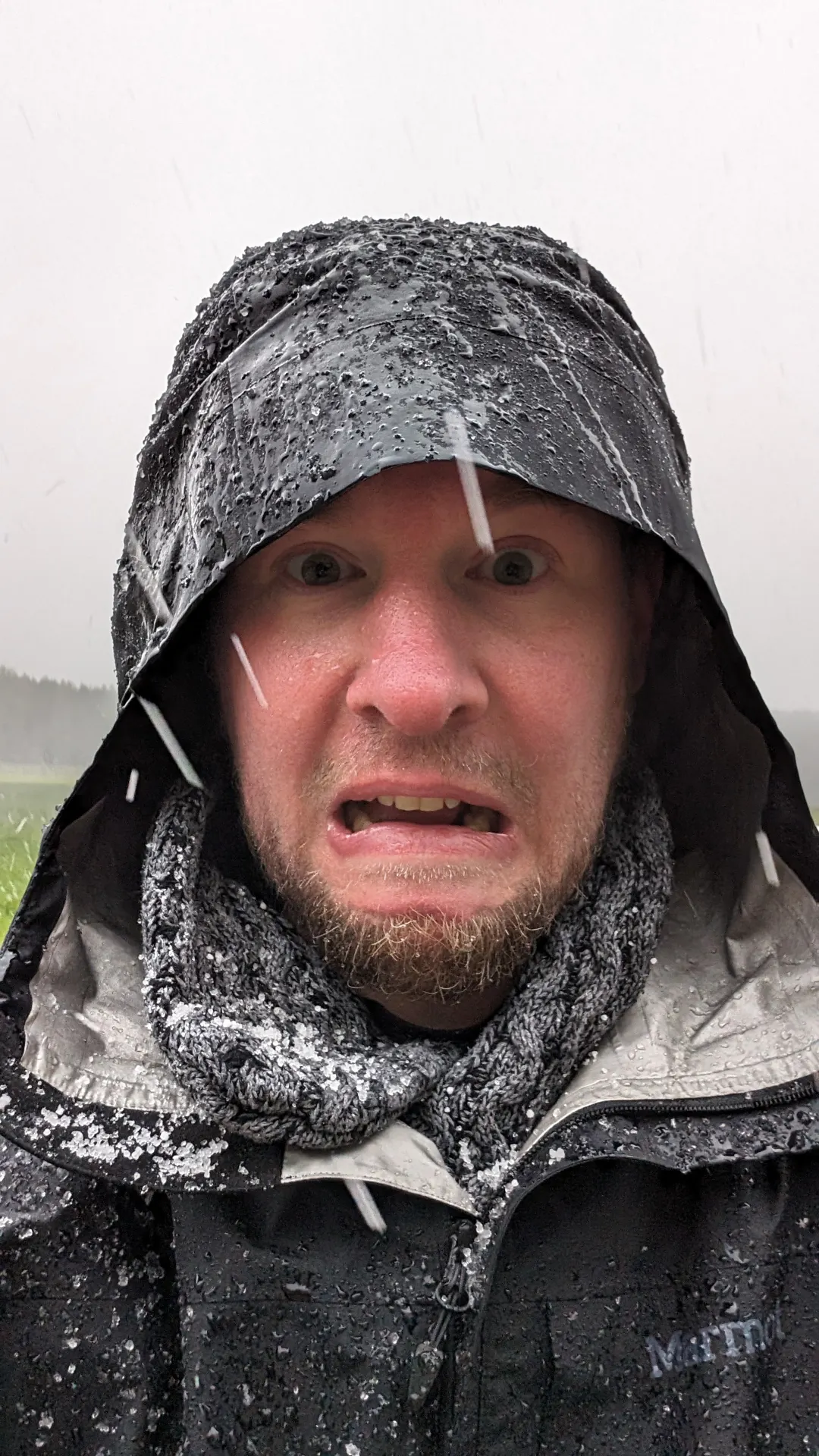 Das Foto zeigt André, Gesichtsausdruck gequält/erschrocken, gehüllt in eine Kapuze, Regen- und Graupeltropfen huschen durchs Bild. Ist das Schnee auf seiner Jacke? Der arme Mann!