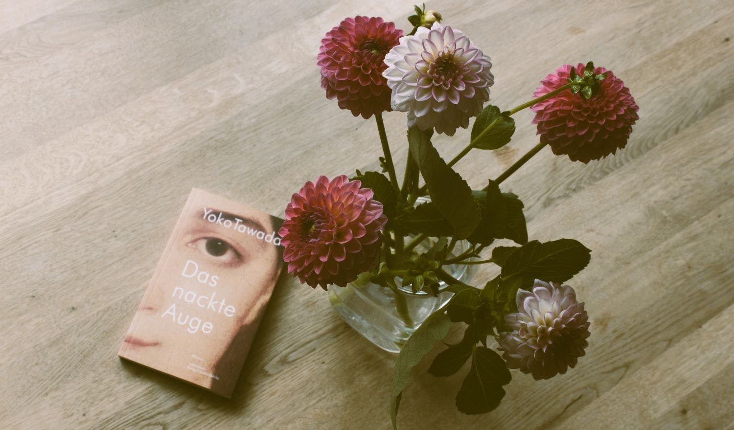 Das Buch "Das nackte Auge" von Yoko Tawada liegt auf einem Tisch. Blumen in einer Vase stehen daneben.
