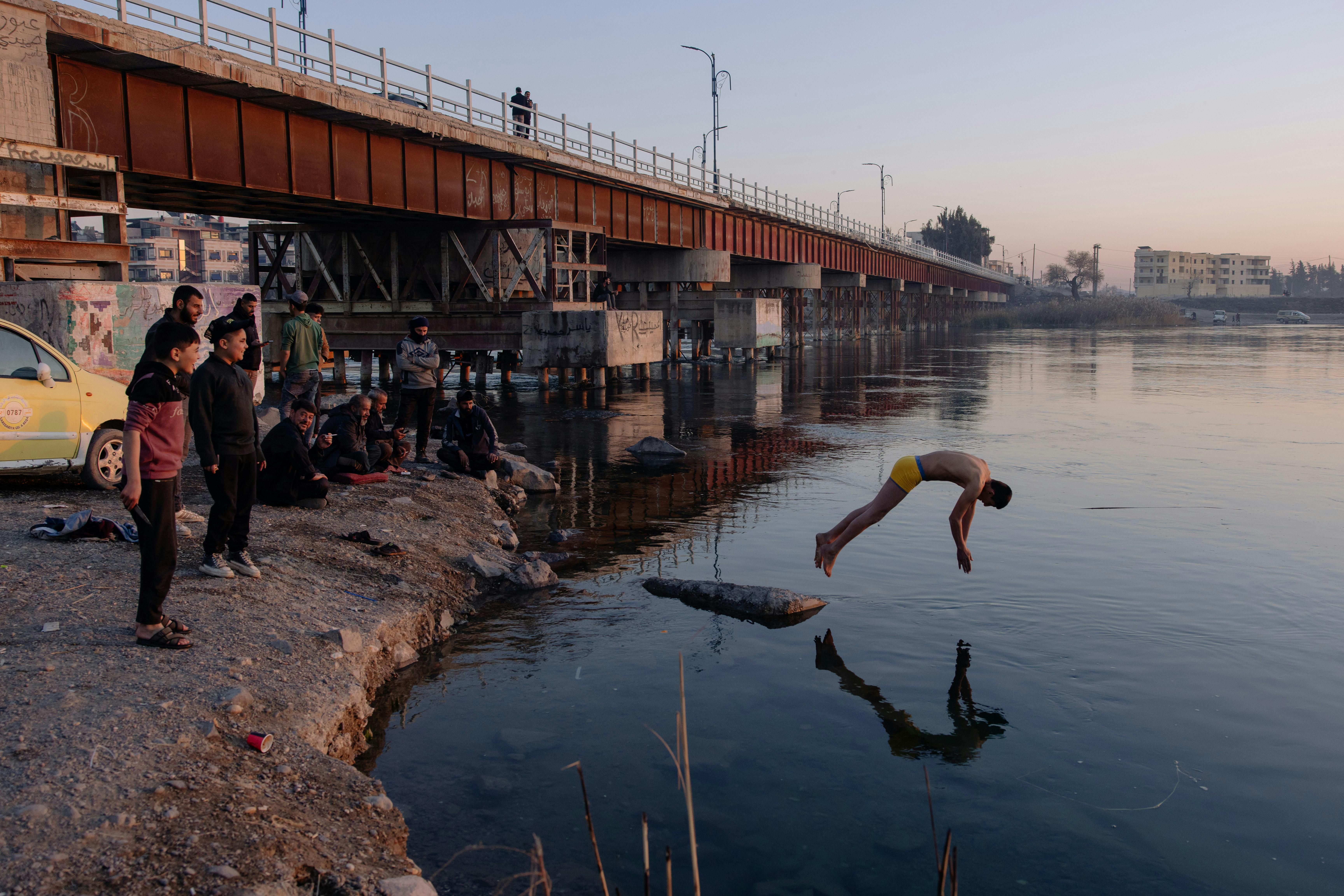 Ein junger Mann springt kopfüber in einen Fluss. Hinter ihm sieht man eine rostige Brücke. Ein paar Kinder schauen zu.