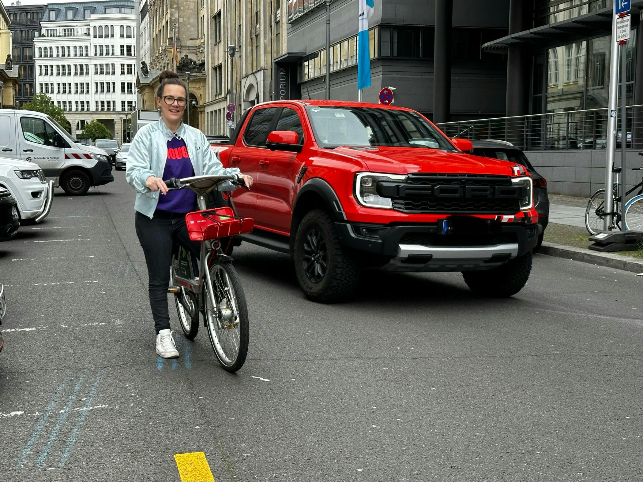 Eine Frau auf einem Fahrrad steht auf einer städtischen Straße und wird von einem riesigen roten Pick Up überholt.