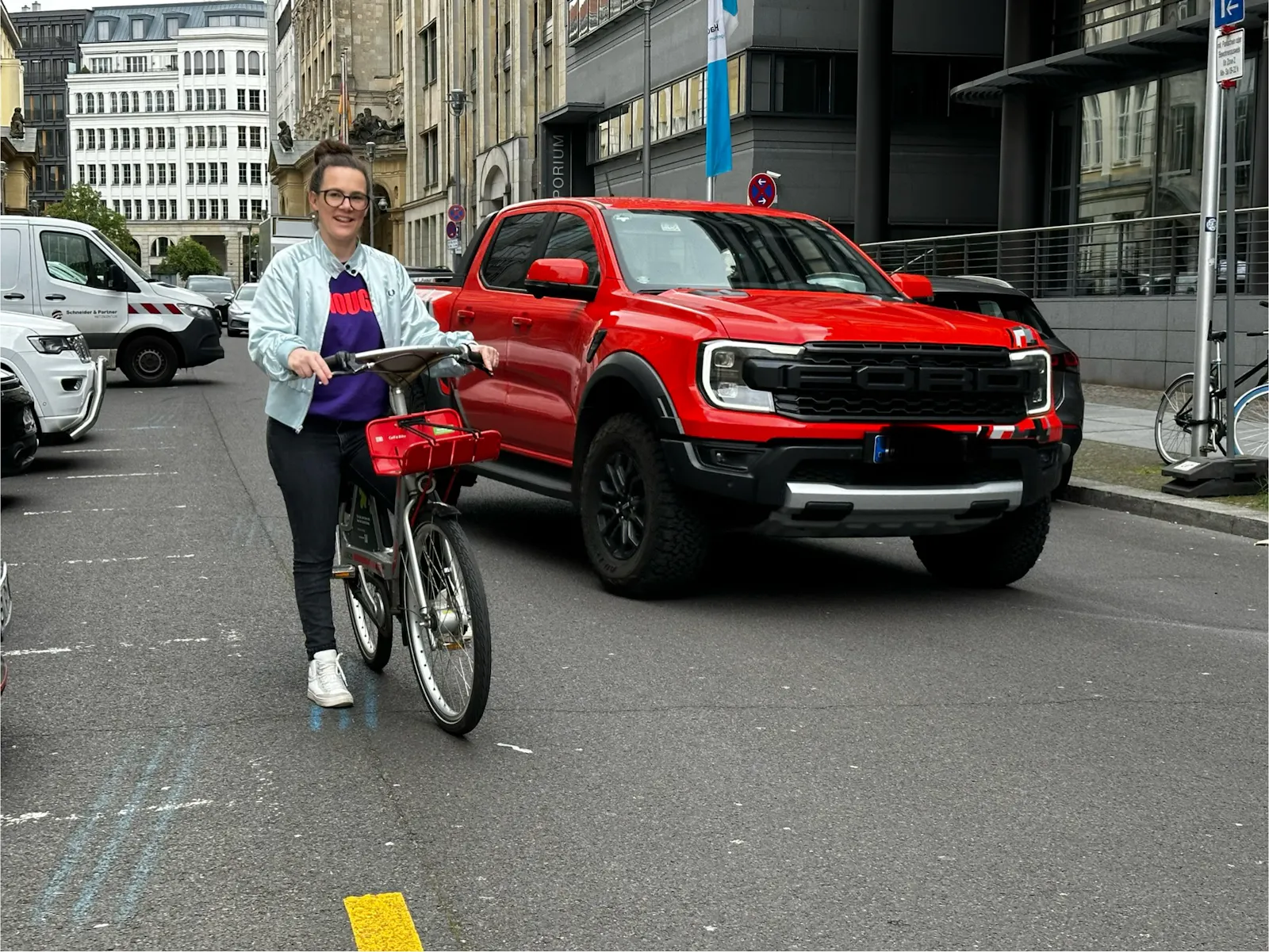 Eine Frau auf einem Fahrrad steht auf einer städtischen Straße und wird von einem riesigen roten Pick Up überholt.