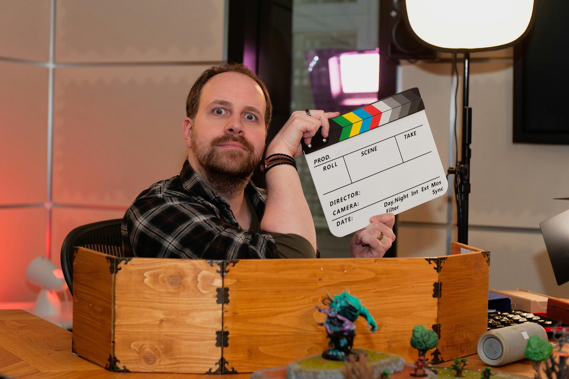 Alex sitzt an seinem DM-Platz und hält eine Filmklappe in der Hand, dabei schaut er mit weit aufgerissenen Augen in die Kamera. Vor seinem DM-Screen stehen einige Miniaturen.