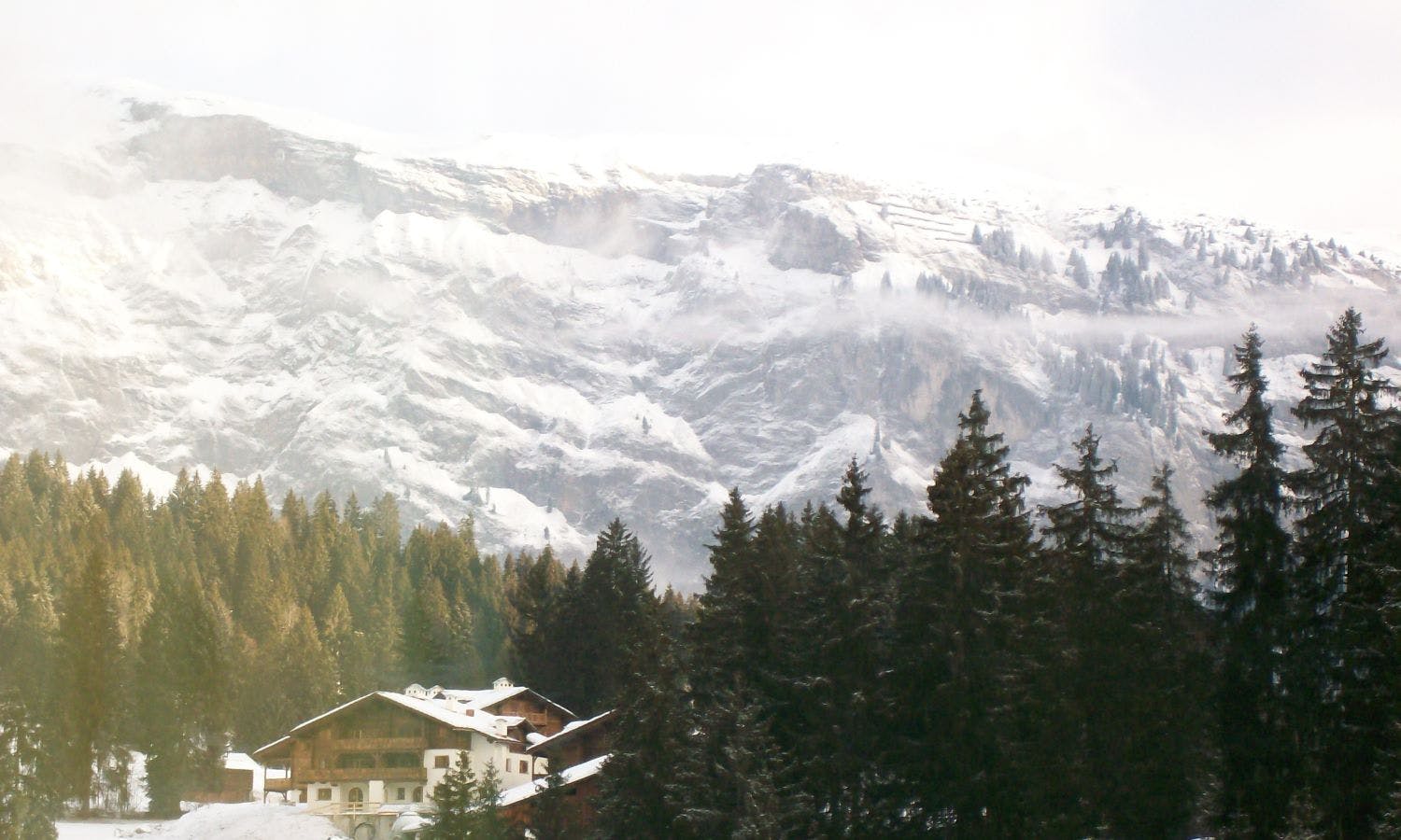 Im Vordergrund ein Haus und dunkle Nadelbäume, im Hintergrund schneebedeckte Berge.