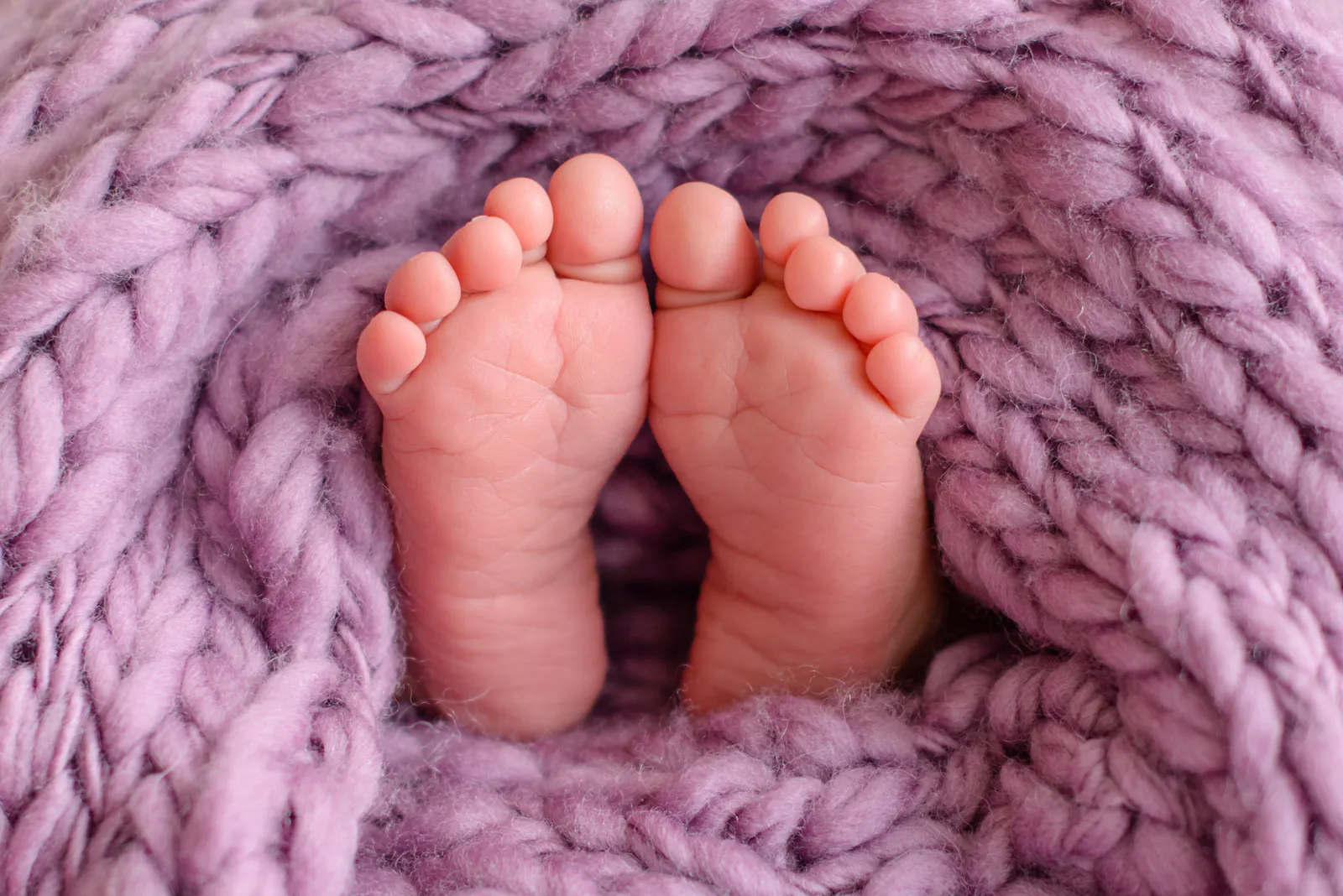 Das Foto zeigt zwei kleine Babyfüße auf einer rosa-lila Strickdecke