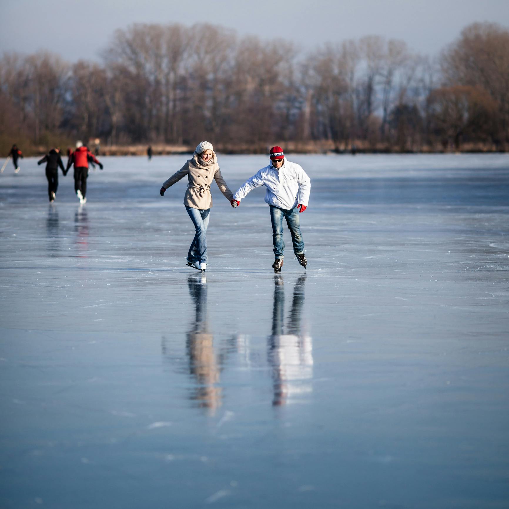 Dieses Foto zeigt ein Paar beim Eislaufen auf einem zugefrorenen See. Im Vordergrund laufen eine Frau und ein Mann Hand in Hand auf dem Eis, im Hintergrund sind weitere Eisläufer und kahle Bäume zu sehen.