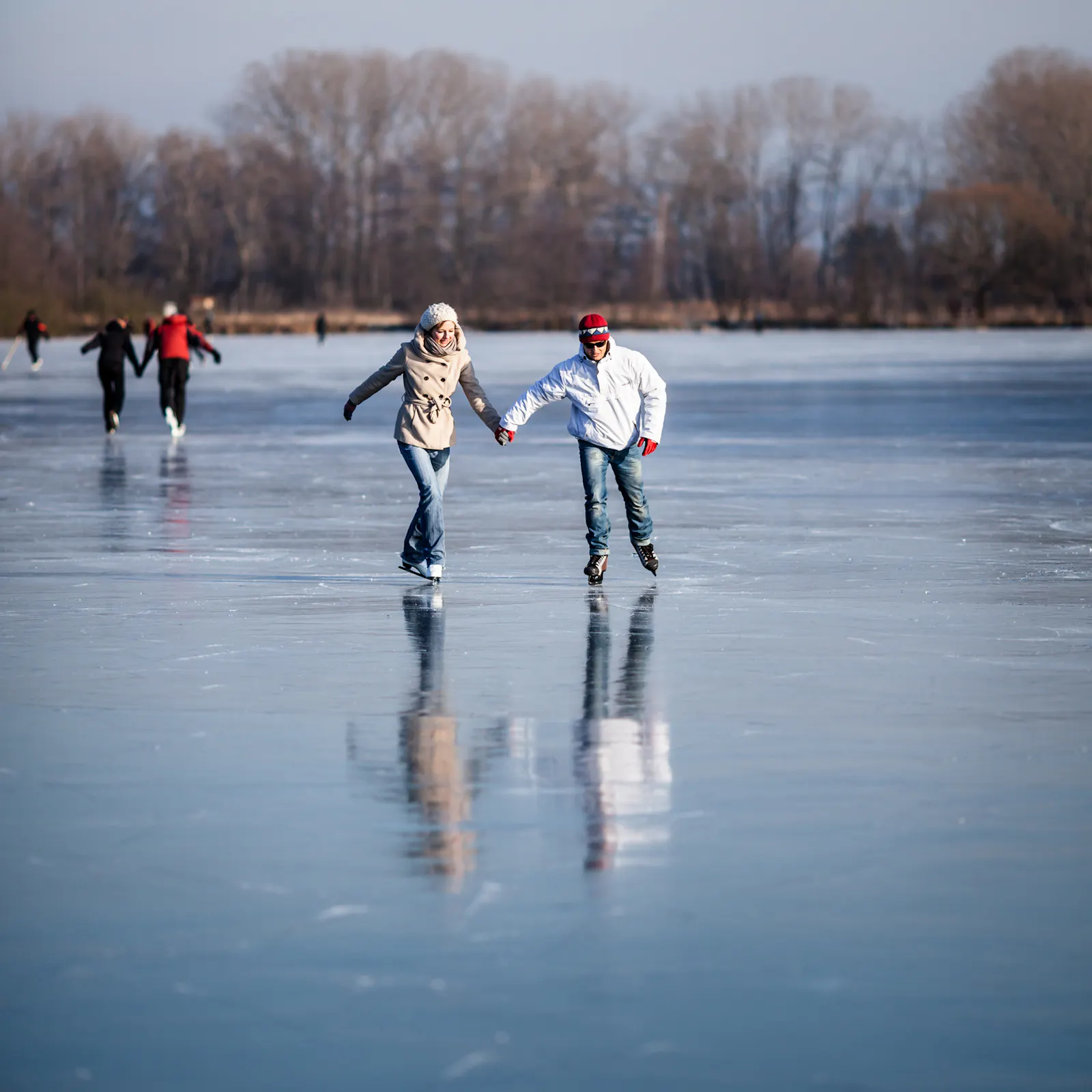 Dieses Foto zeigt ein Paar beim Eislaufen auf einem zugefrorenen See. Im Vordergrund laufen eine Frau und ein Mann Hand in Hand auf dem Eis, im Hintergrund sind weitere Eisläufer und kahle Bäume zu sehen.