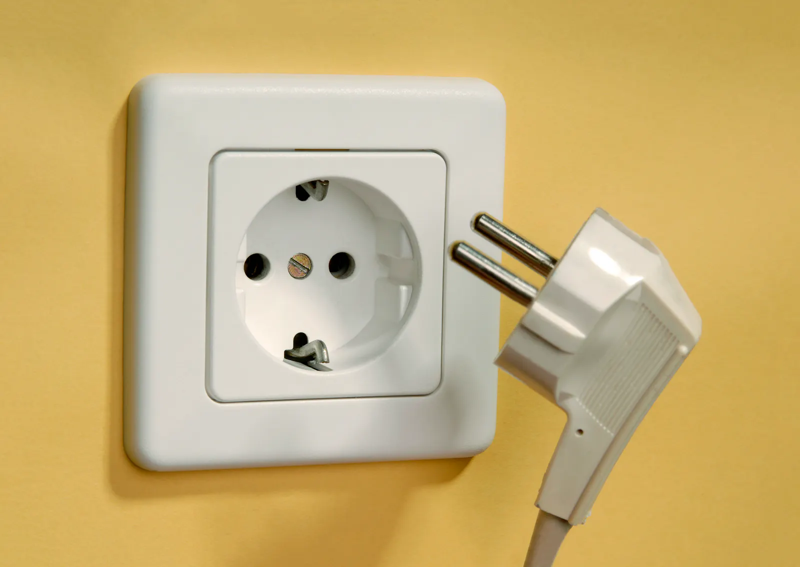 Das Bild zeigt eine weiße Steckdose an einer gelben Wand. Daneben hängt ein weißer Stecker, der noch nicht eingesteckt ist.