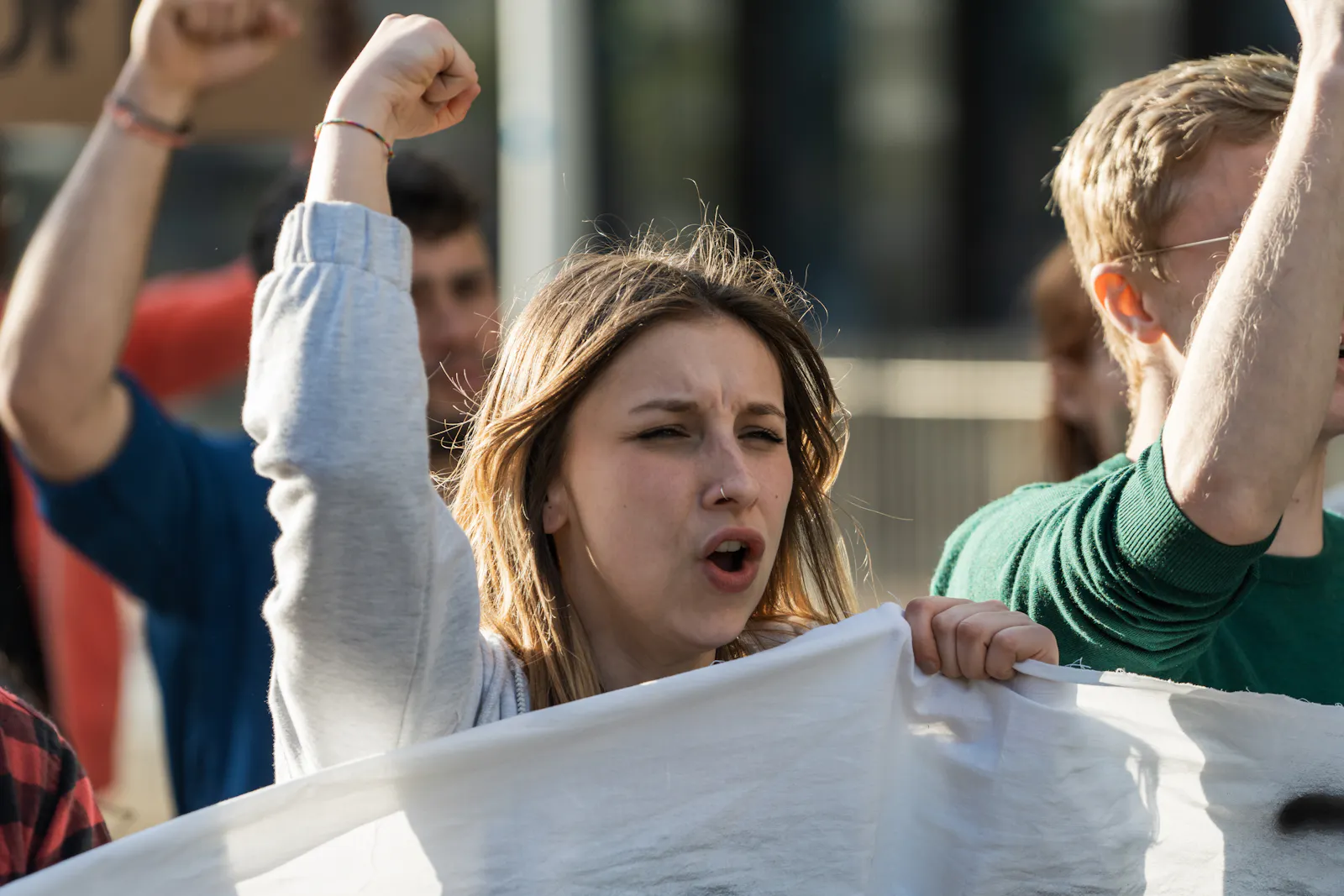 Das Bild zeigt eine junge Frau bei einer Demonstration. Sie hält die Faust in die Luft und ruft etwas. Im Hintergrund sind weitere Demonstrierende zu sehen.