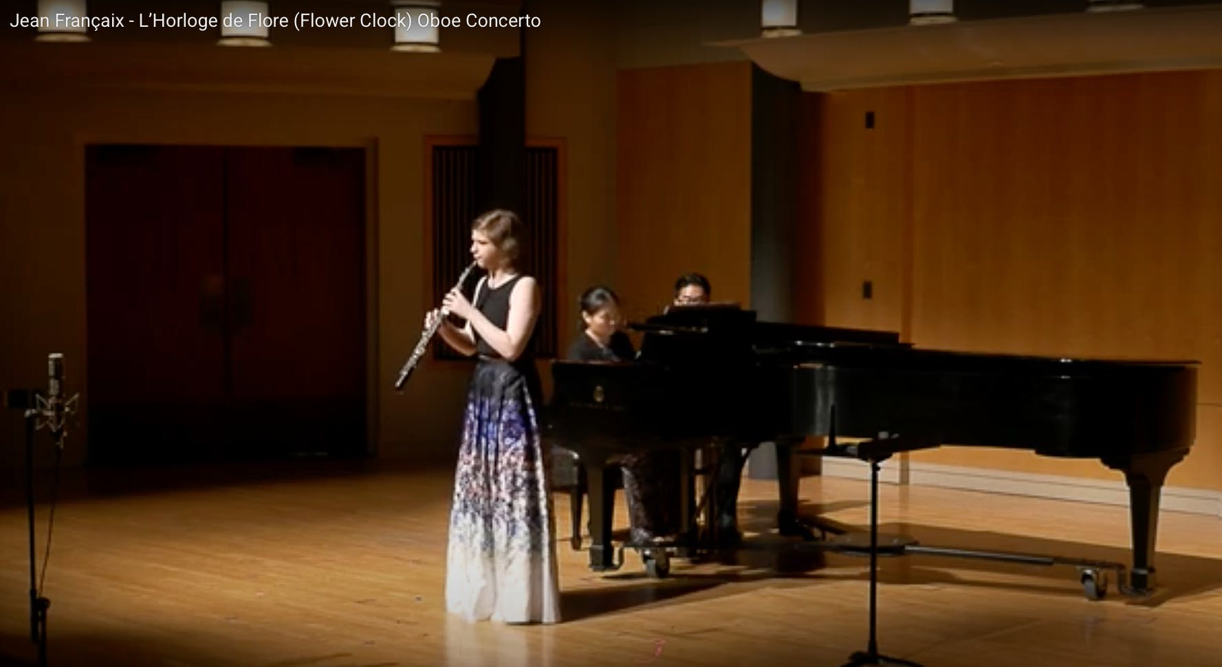 Eine junge Frau mit einem langen Kleid steht vor einem Flügel und spielt auf einer Oboe.