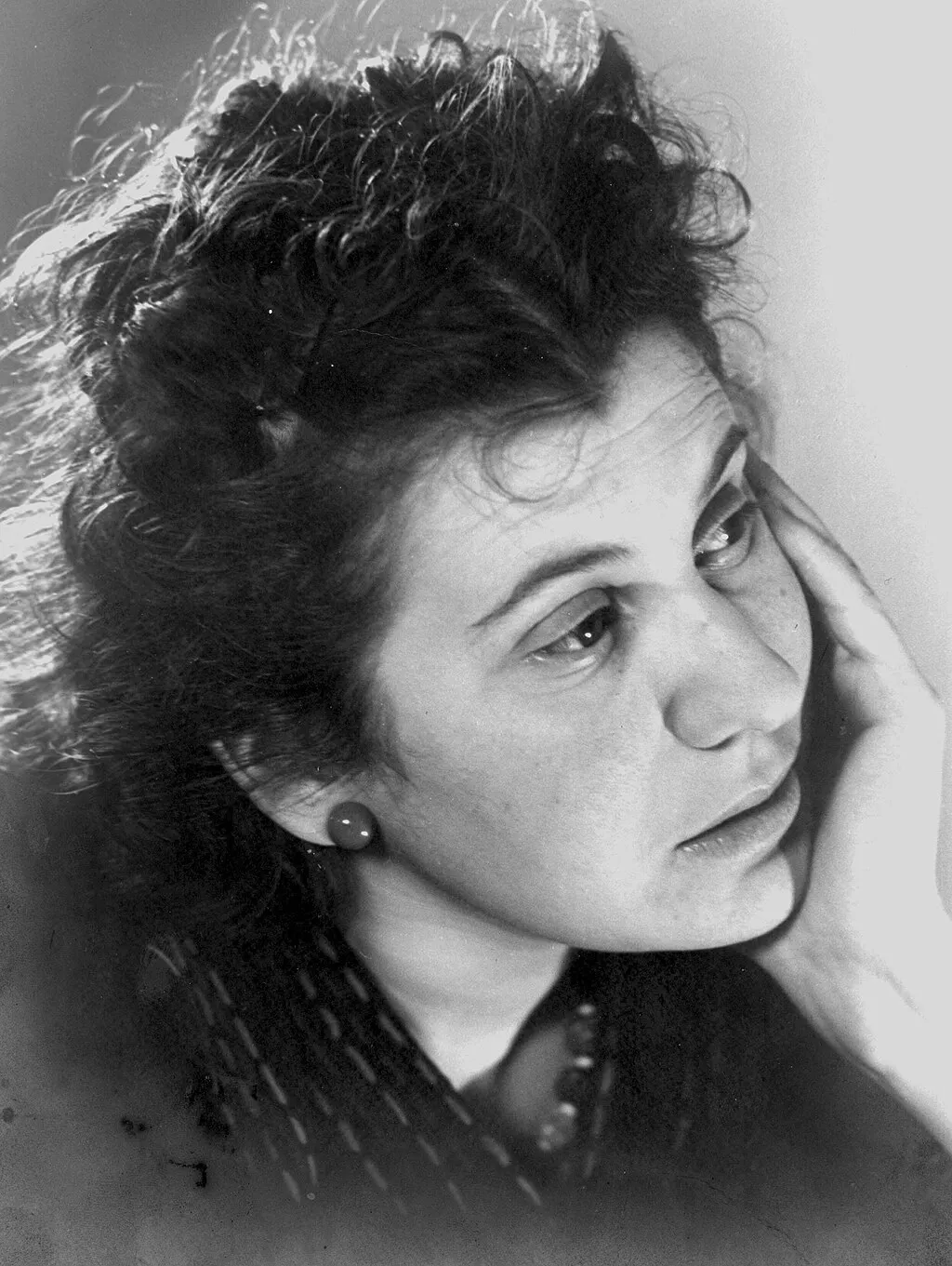 Ein schwarz-weiß Foto einer jungen Frau mit dunklen haaren, die ihr Kinn gestützt in der Hand hält.