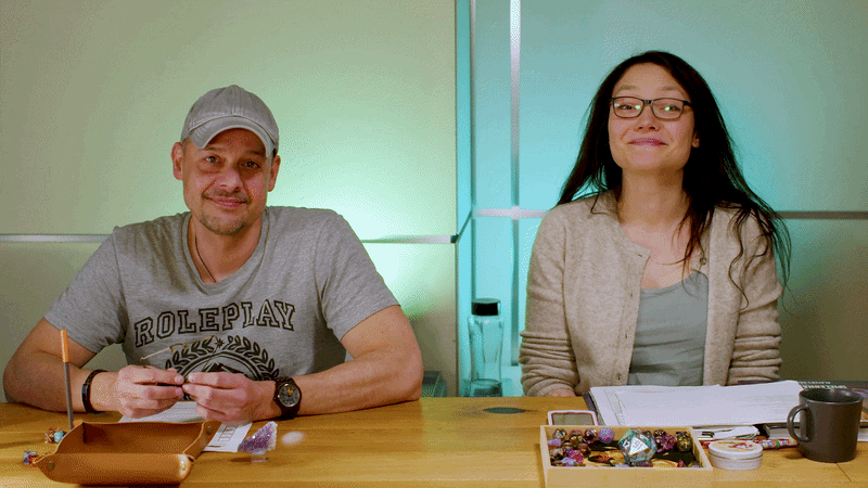 Marcel und Rubi sitzen nebeneinander an unserem Spieltisch vor einer türkis beleuchteten Wand und lächeln und winken in die Kamera. Links sitzt Marcel mit grauem Cap und einem grauen „Roleplay"-T-Shirt, vor ihm liegt ein Dice Tray mit ein paar Würfeln drum herum. Rechts sitzt Rubi mit Brille und langem dunklen Haar, einem beigen Cardigan, vor ihr liegen Notizblätter, eine offene Würfelbox mit bunten Würfeln und eine dunkle Tasse.