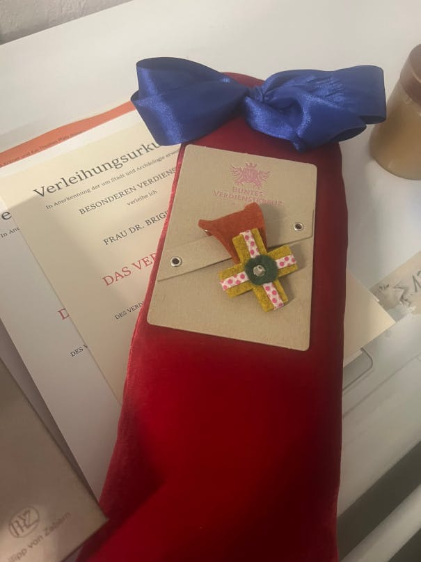 Foto zeigt ein rotes langes Kissen auf dem ein Orden in Form eines Kreuzes aus buntem Filz liegt. Darunter eine Verleihungsurkunde im Stil der offiziellen Urkunden der Bundesrepublik.