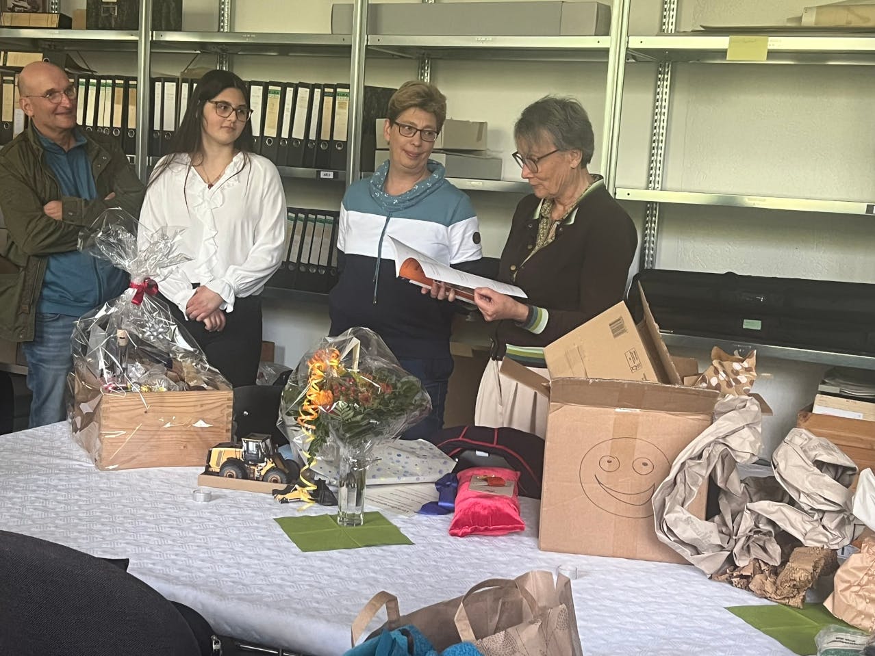 Brigitta Kunz steht neben einen anderen Personen hinter einem Tisch, mit Geschenken und Verpackungspapier. Sie blättert in einem Buch, die überreichte Festschrift Kunz.