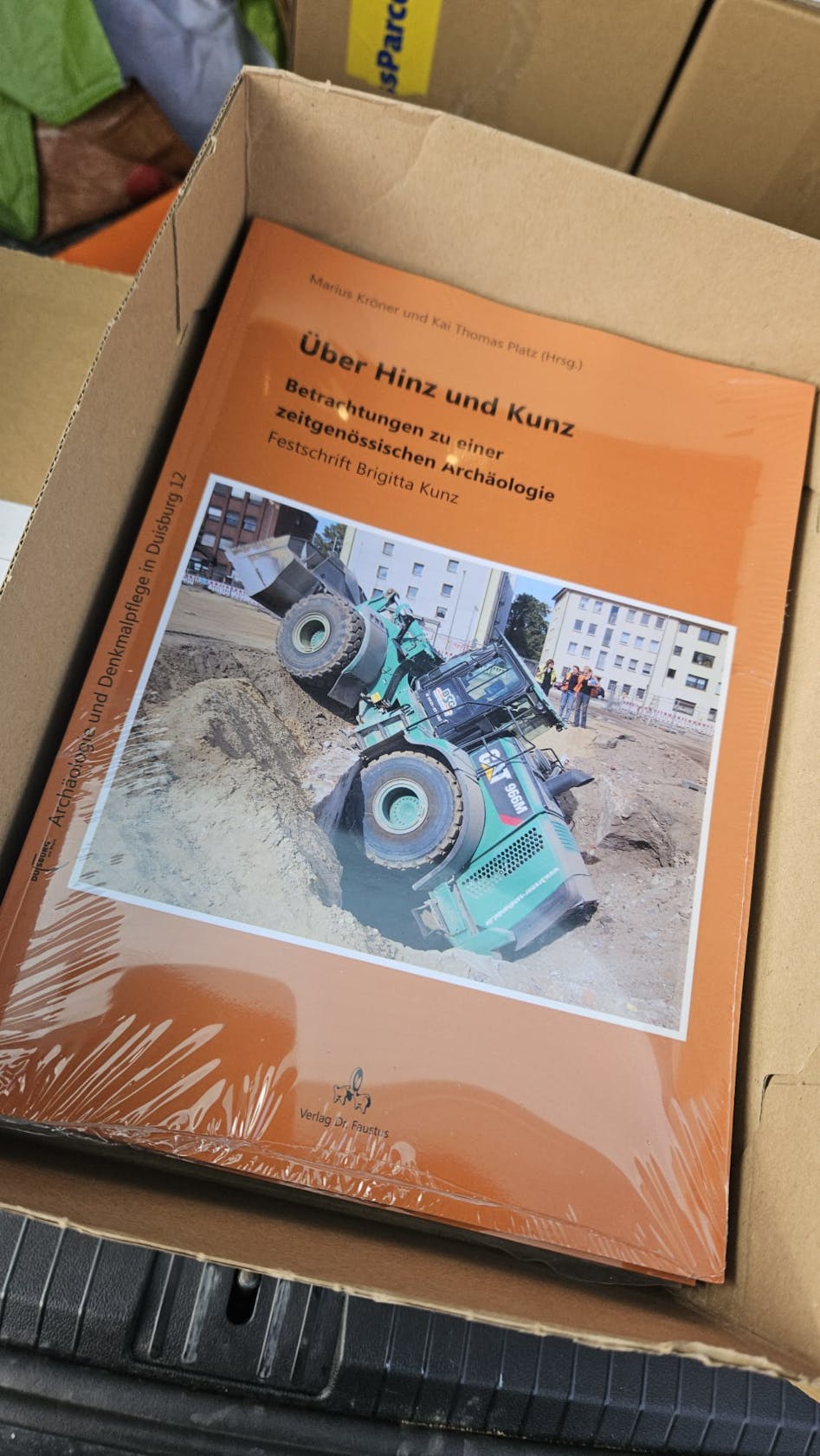 Die Festschrift in klare Folie eingepackt in einem Karton. Titelbild der Festschrift Brigitta Kunz. Das Titelblatt ist orange und in der Mitte ist ein Foto mit einem grünen Radlader, der in einen Grabungsschnitt gekippt ist.