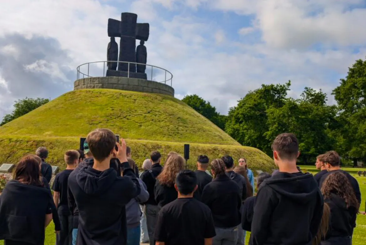 Eine Schülergruppe vor einem Denkmal der deutschen Kriegsgräberstätte auf einem Friedhof in La Cambe. Das dunkle Kreuz mit menschlichen Figuren darunter steht auf einem künstlichen mit Gras bewachsenen Hügel.