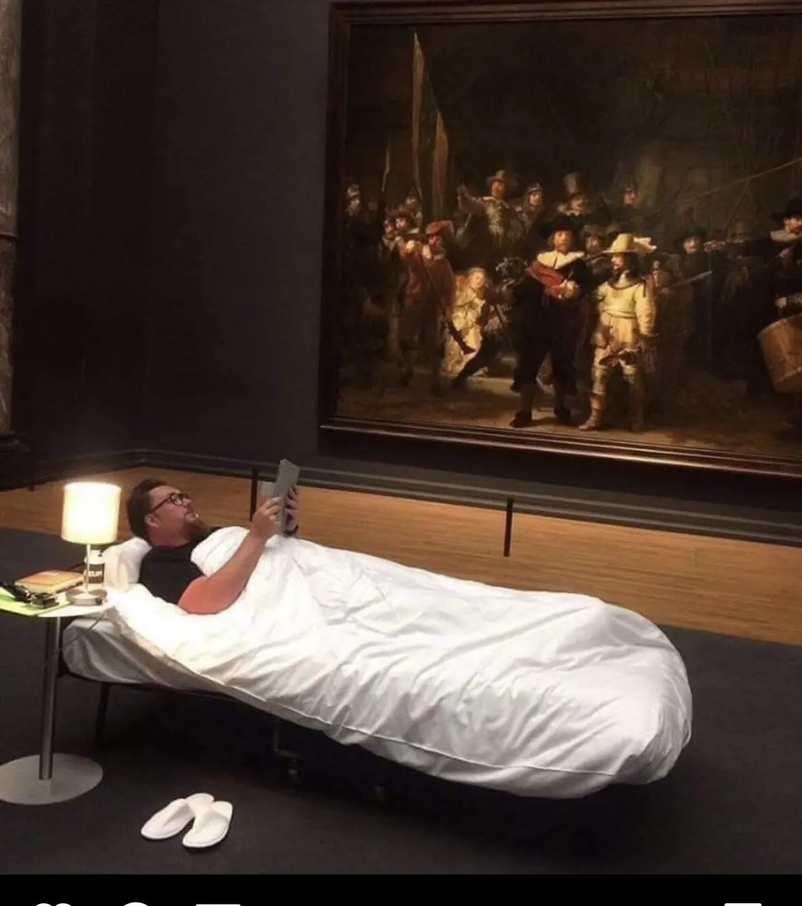 Mensch liegt im Museum mit Buch im Bett vor Gemälde