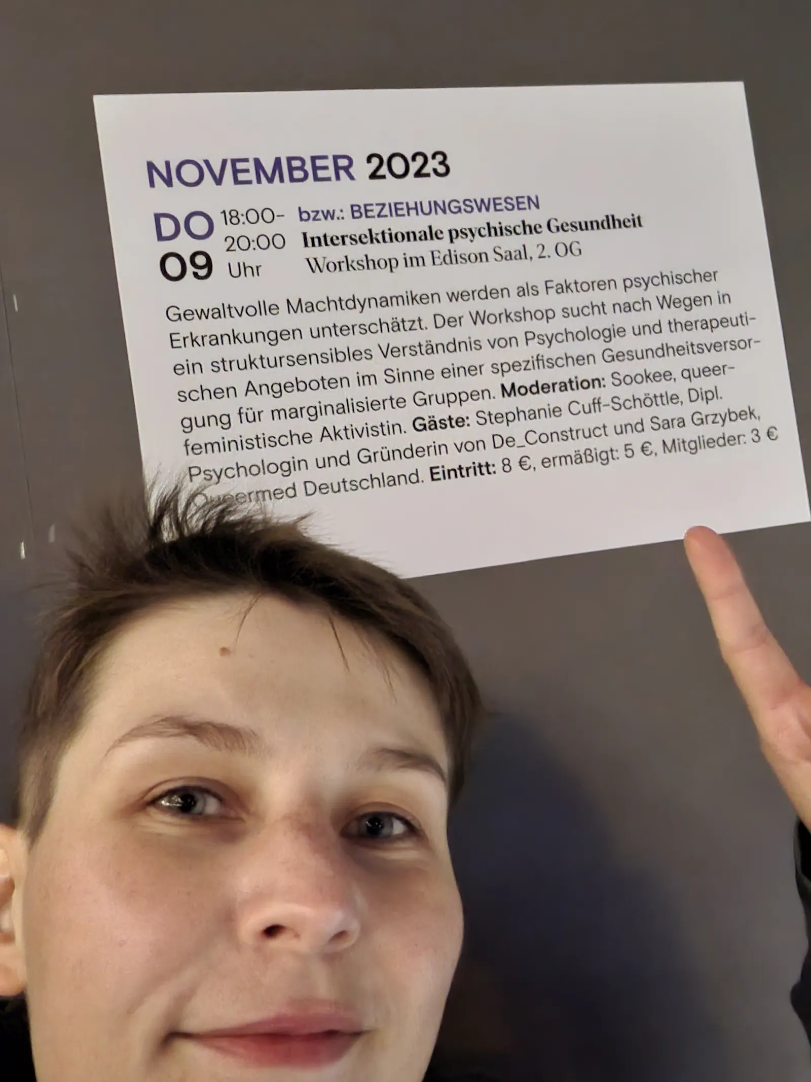 Queermed Gründer*in Sara Grzybek zeigt mit einem Zeigefinger auf ein Veranstaltungsposter. Die Veranstaltung lautet "Intersektionale psychische Gesundheit" und fand am 09.11.2023 im Urania in Berlin statt.