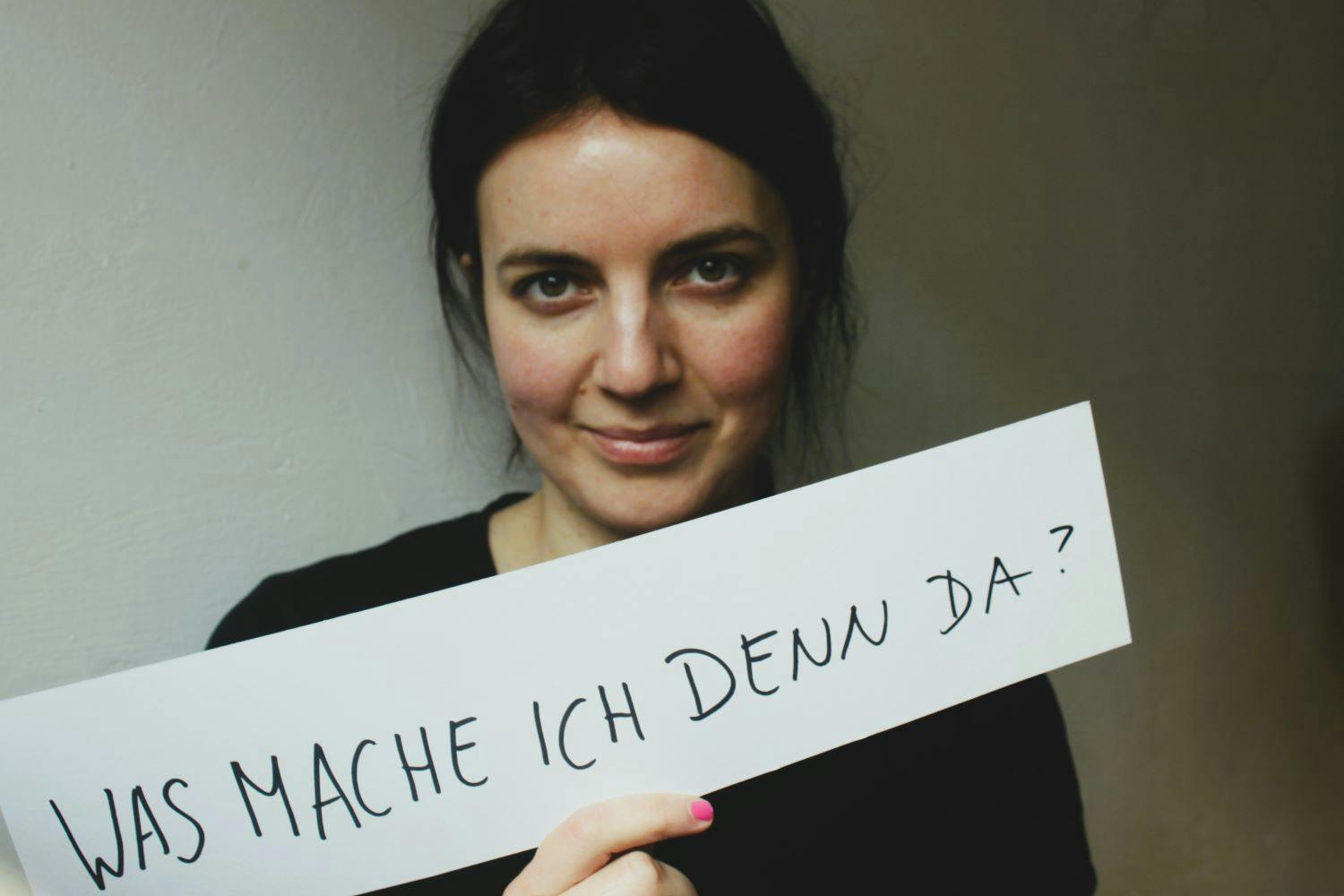 Eine Frau schaut frontal in die Kamera und hält ein weißes Schild mit der Aufschrift: Was mache ich denn da?