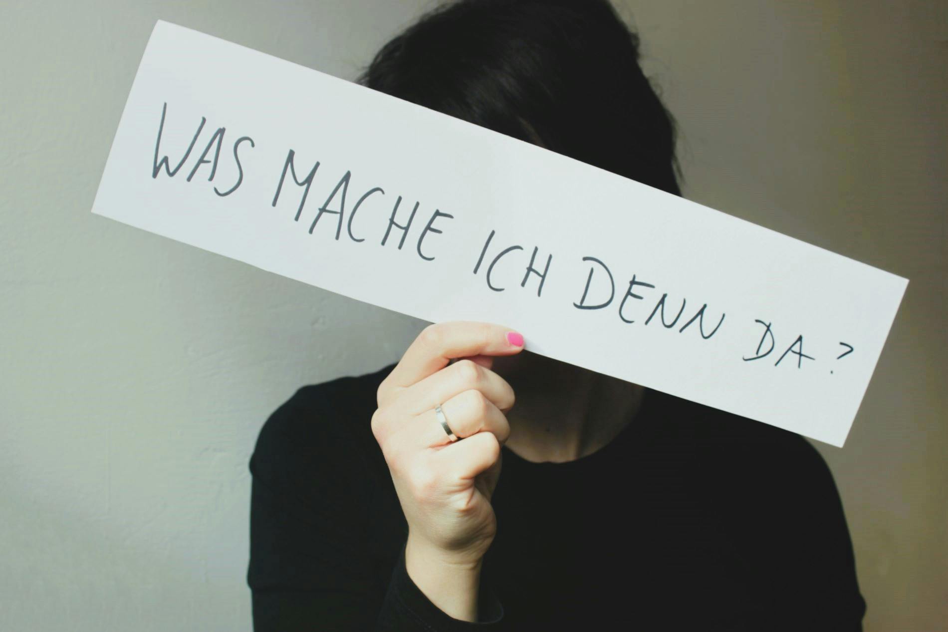 Eine Person  hält ein weißes Schild in die Kamera, auf dem "Was mache ich denn da?" steht.