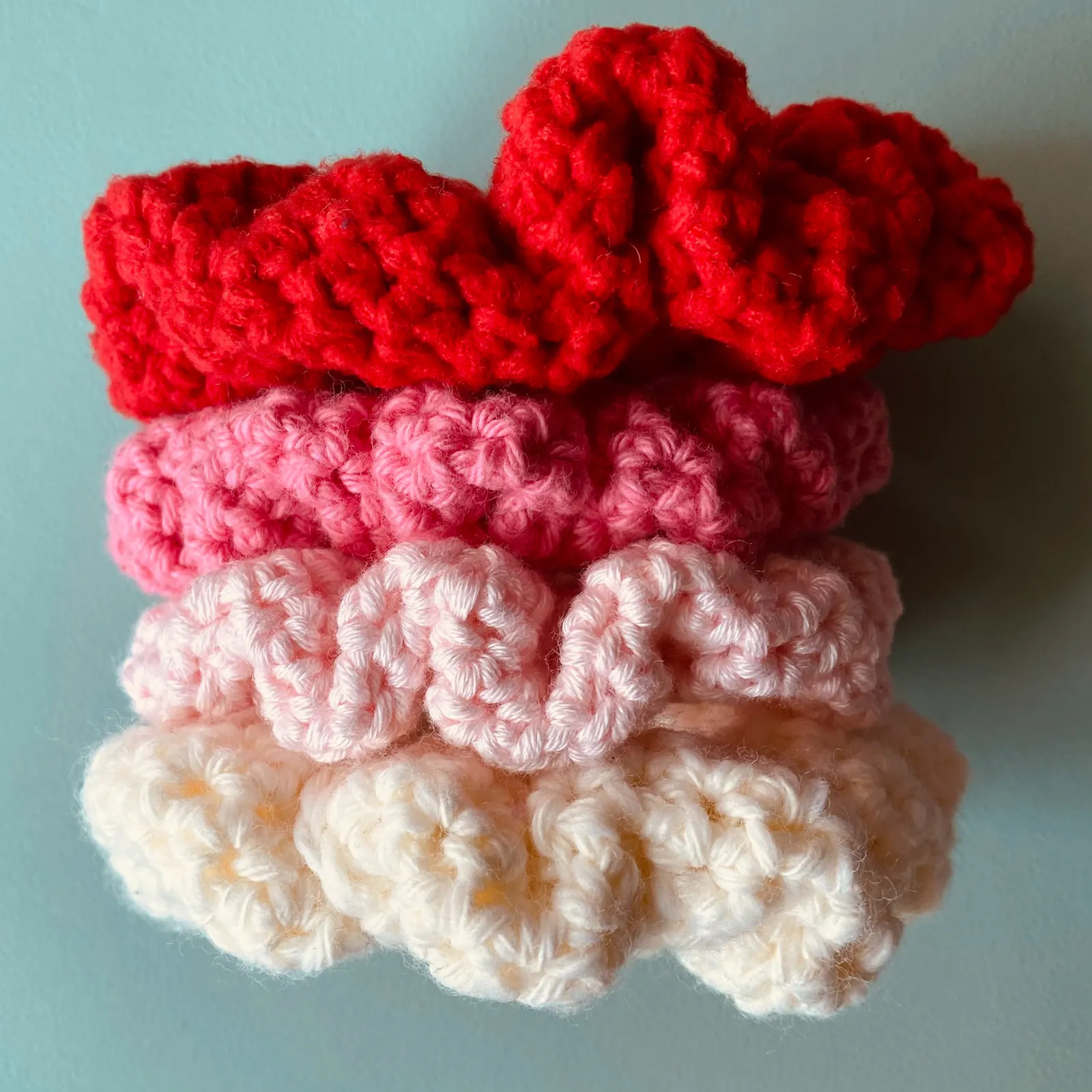Vier verschiedenfarbige Scrunchies übereinander gestapelt. Die Haargummis sind cremfarben (unten), hellrosa, dunkelrosa und rot (oben) und scheinen gehäkelt worden zu sein.