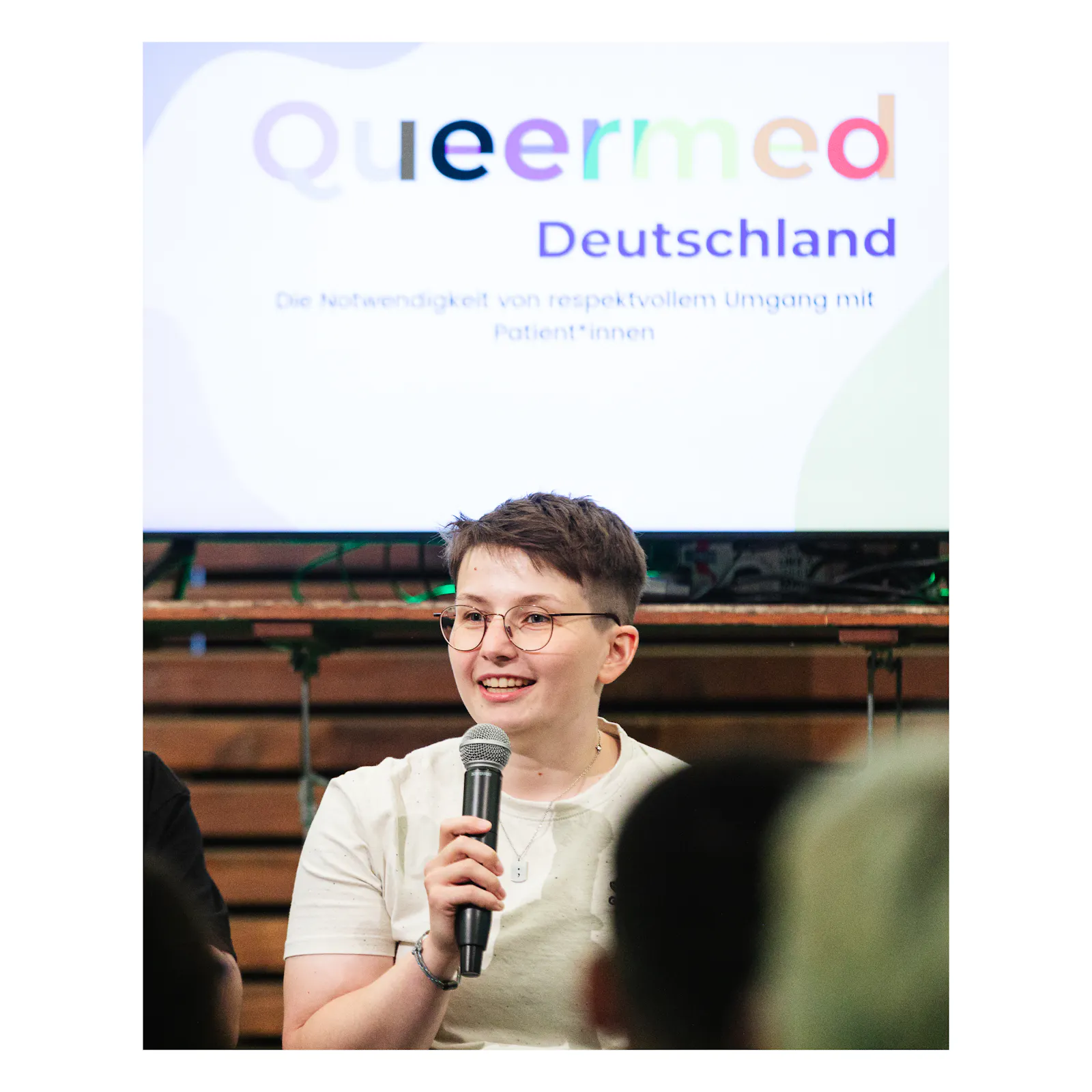 Queermed Gründer*in Sara Grzybek hält ein Mikrofon in der Hand und spricht vor einem Publikum. Im Hintergrund ist auf einem Fernseher der erste Slide einer Präsentation zu sehen.