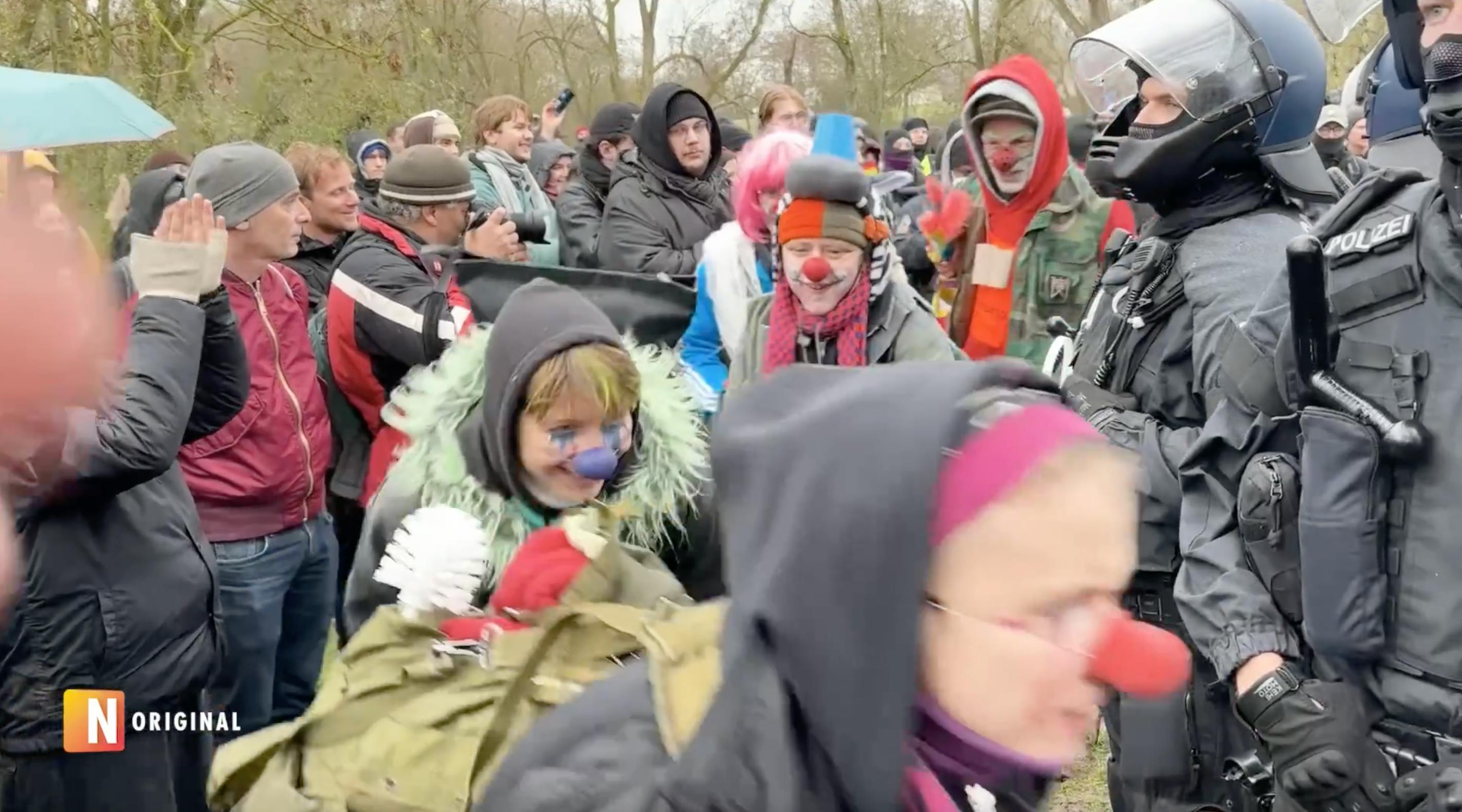 Menschen mit Clownsnasen tanzen im Kreis um von schwer gepanzerten Polizisten geschützte Nazis