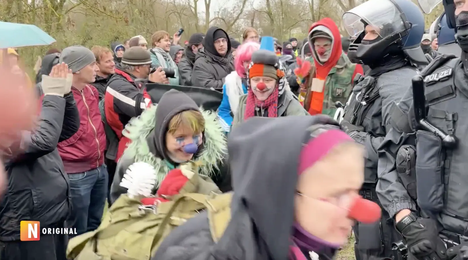 Menschen mit Clownsnasen tanzen im Kreis um von schwer gepanzerten Polizisten geschützte Nazis