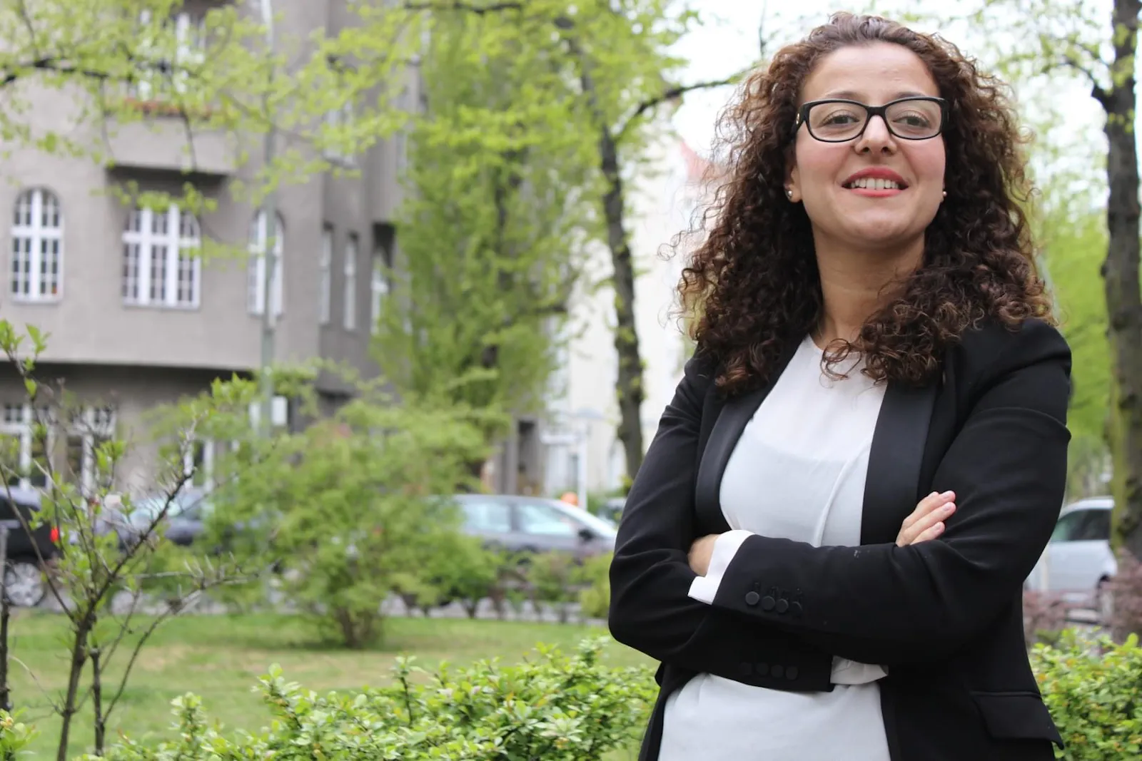 Jouanna Hassoun steht draußen auf der Straße. Sie trägt ein weißes Shirt und einen schwarzen Blazer und lächelt in die Kamera.