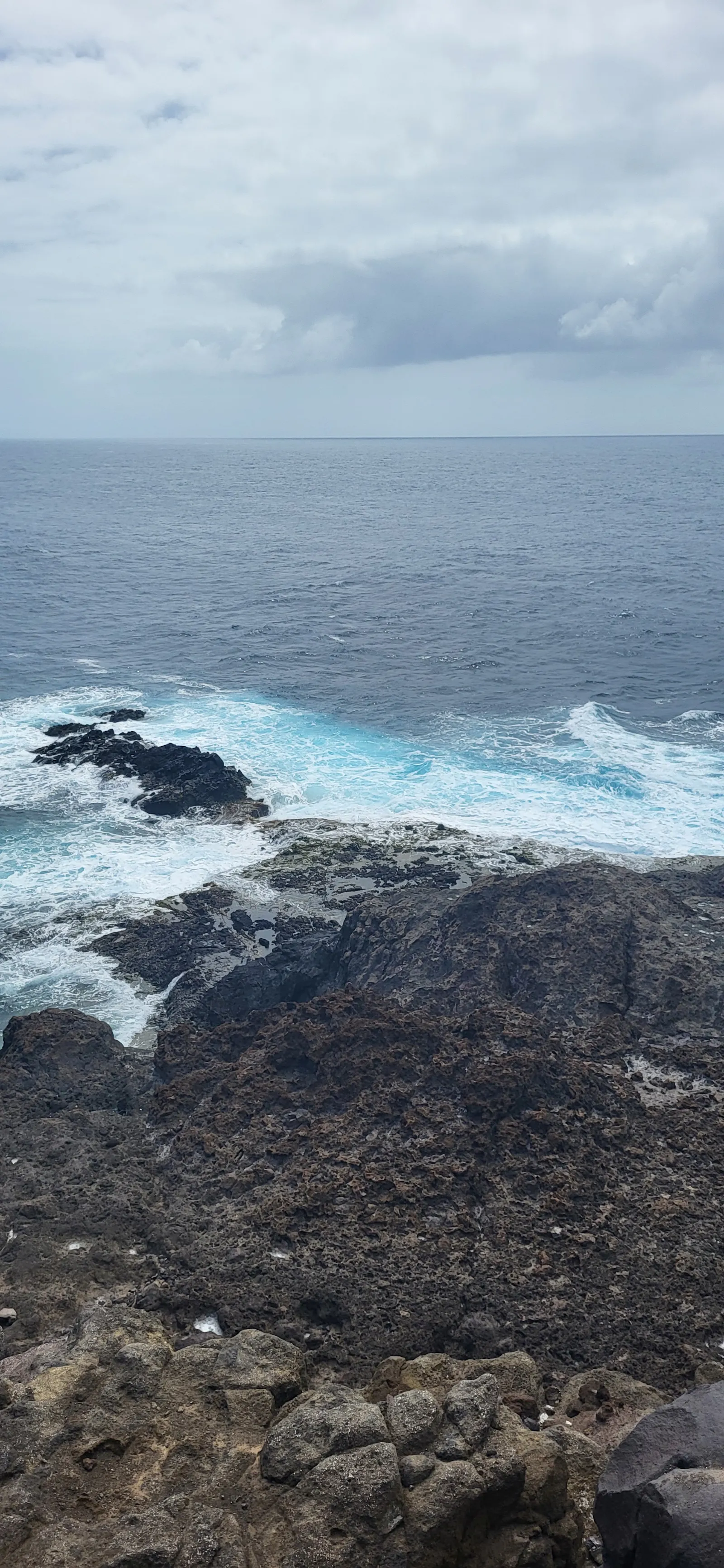Blick auf das Meer von einem Vulkanischen Hang aus. Darüber ein Wolkiger, aber heller Himmel. Wo das Meer auf das felsige Land trifft, ist es aufgewühlt
