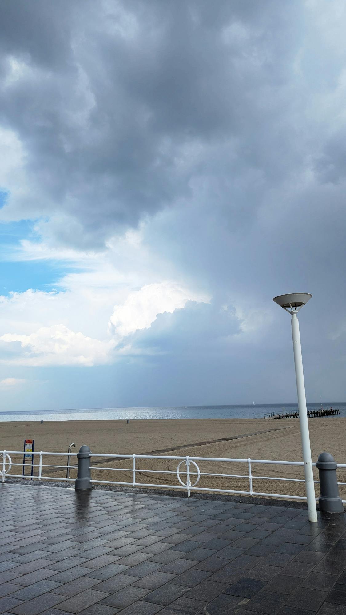 Regennasses Pflaster einer Strandpromenade, dahinter Strand, dahinter Meer, über allem dunkelgraue und leuchtend weiße Wolken und ein Fleck blauer Himmel.